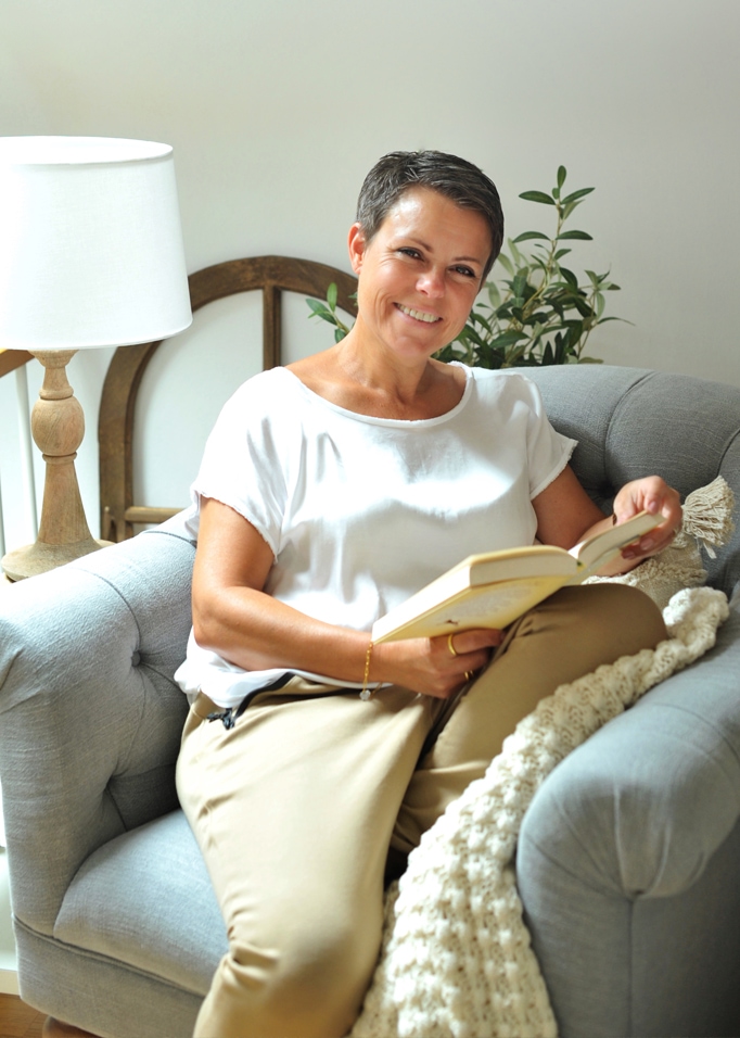 Donna che legge un libro su un divano grigio con coperta bianca e lampada accanto