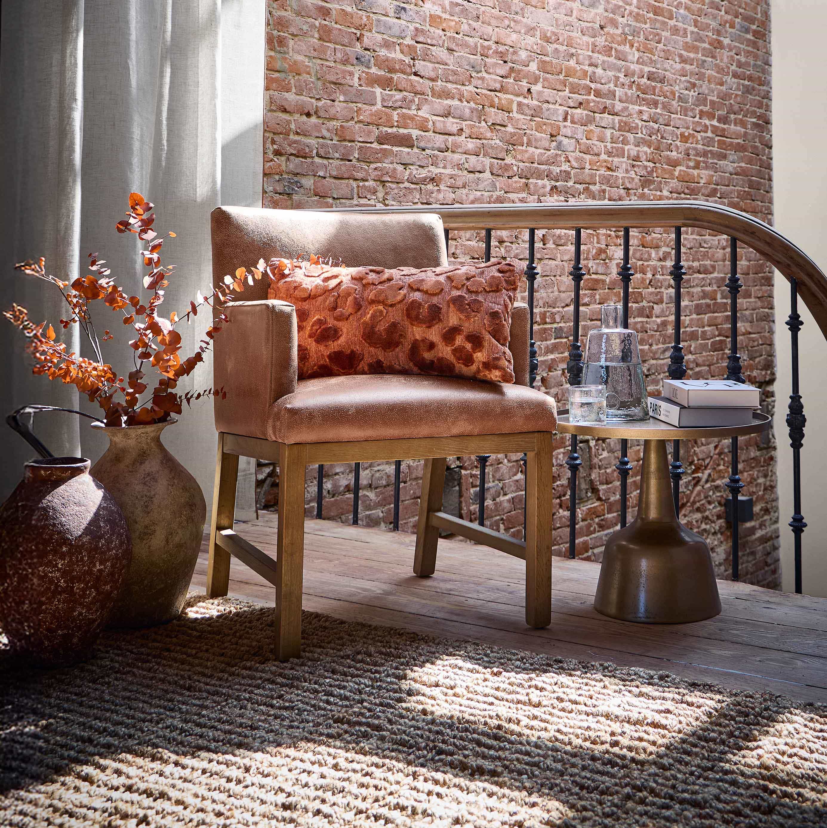 Poltrona beige con base in legno e cuscino arancione davanti a muro di mattoni e tende