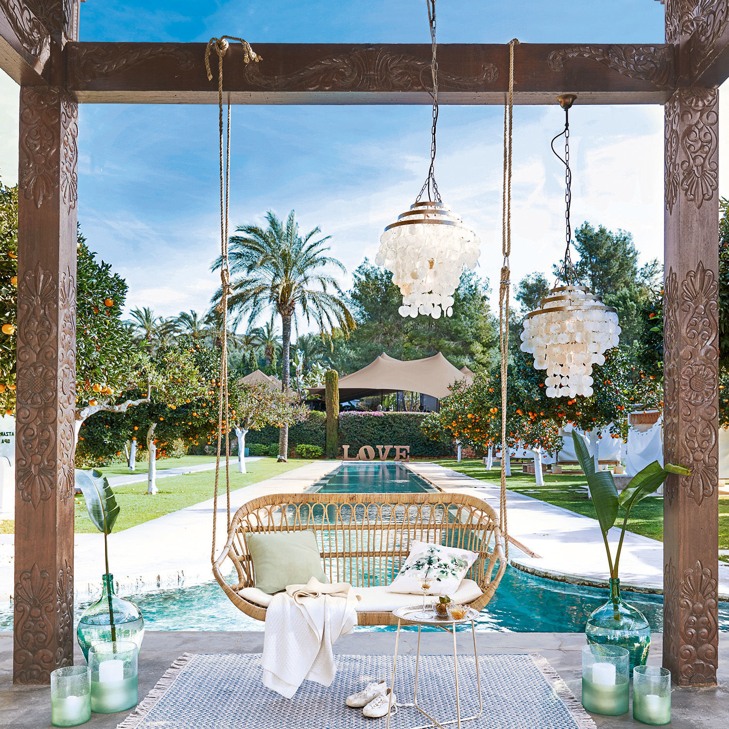 Dondolo sospeso William Altalena in rattan con cuscini e coperta vicino a piscina in giardino con palme e aranci