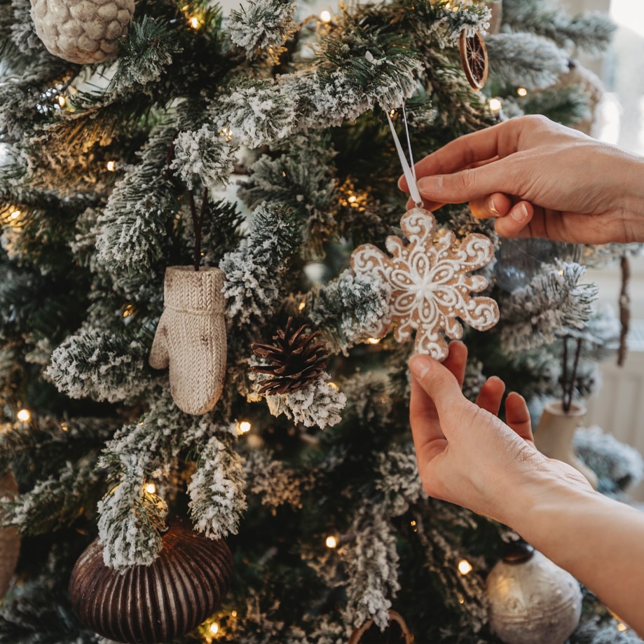 Mani che appendono ornamento a forma di fiocco di neve su albero di Natale innevato con luci