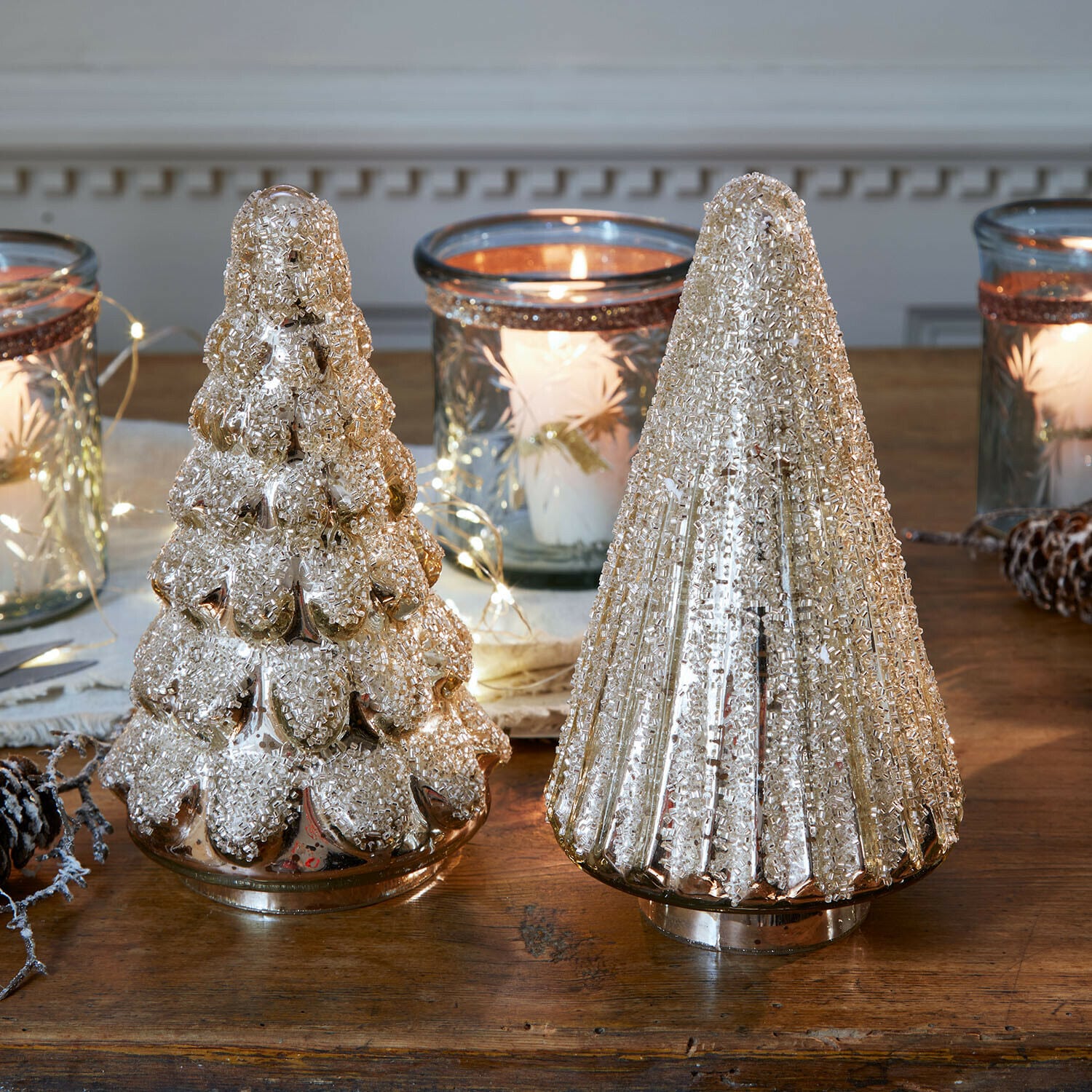 Set di 2 alberi decorativi Domélys Due decorazioni natalizie a forma di albero in vetro dorato con texture, su tavolo in legno con lanterne e candele accese