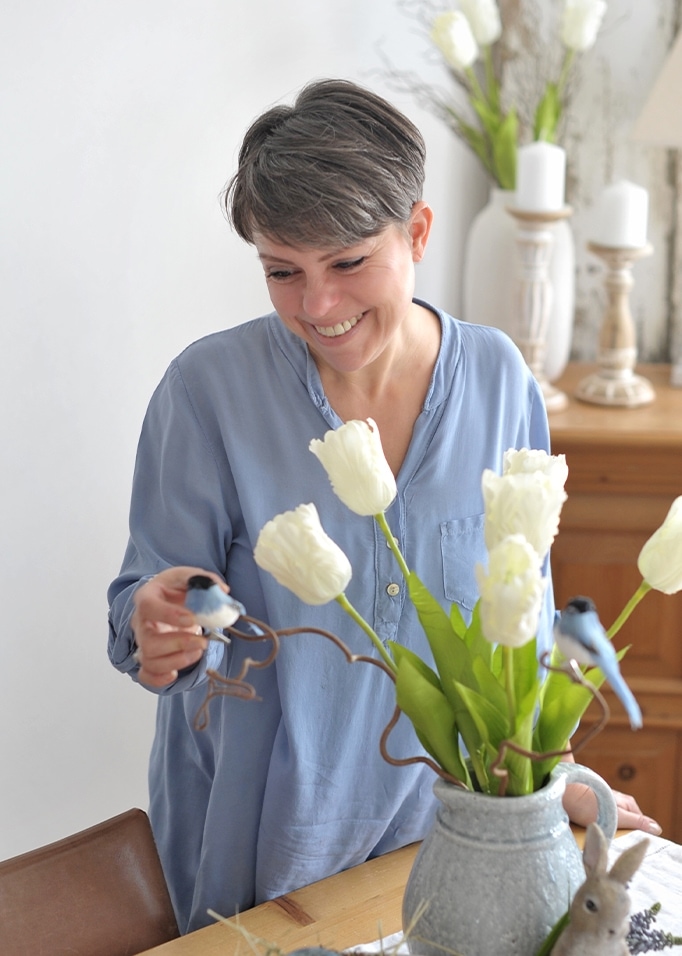 Donna con camicia azzurra decora tavolo con vaso in ceramica grigia, tulipani bianchi e uccelli decorativi