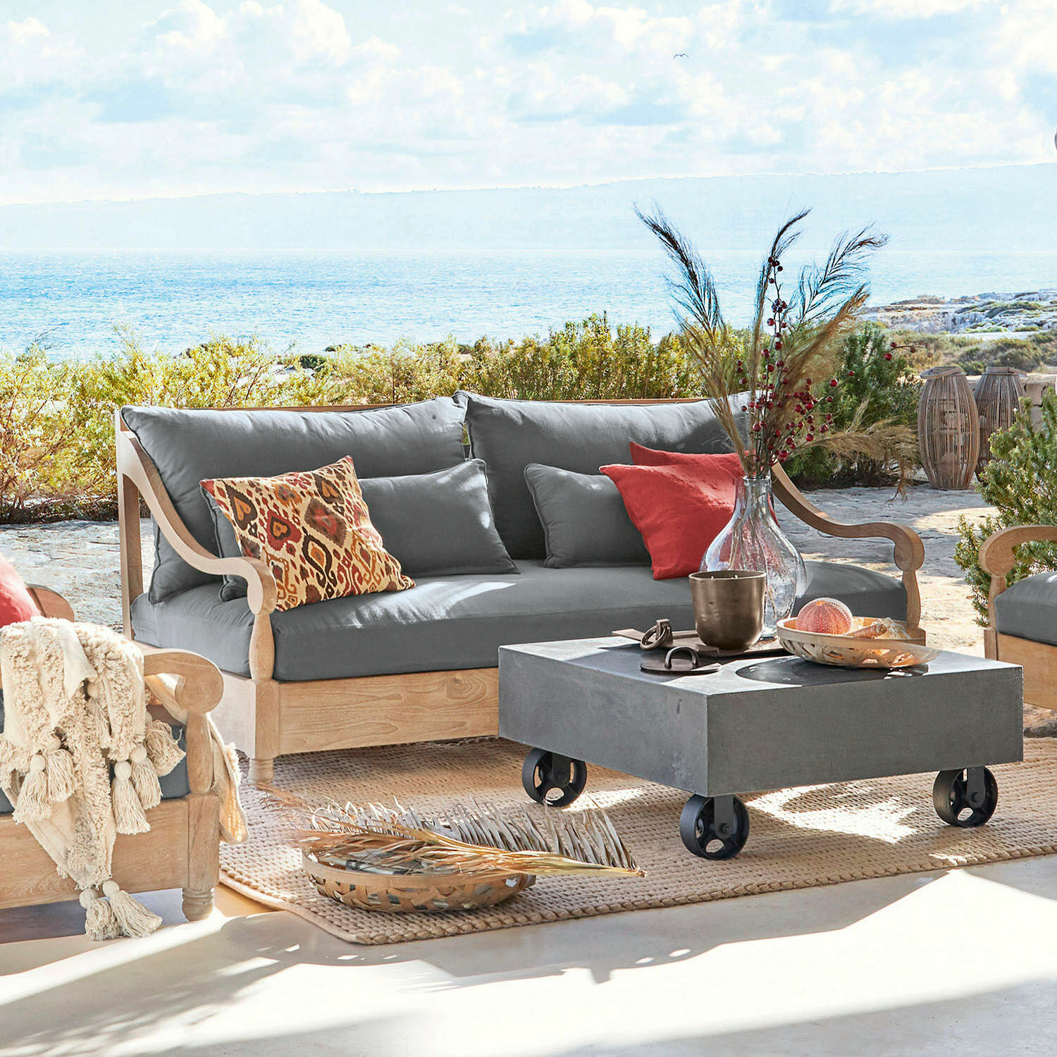 Divano Montcassin Divano da esterno in legno chiaro con cuscini grigi e cuscini decorativi colorati su terrazza con vista mare