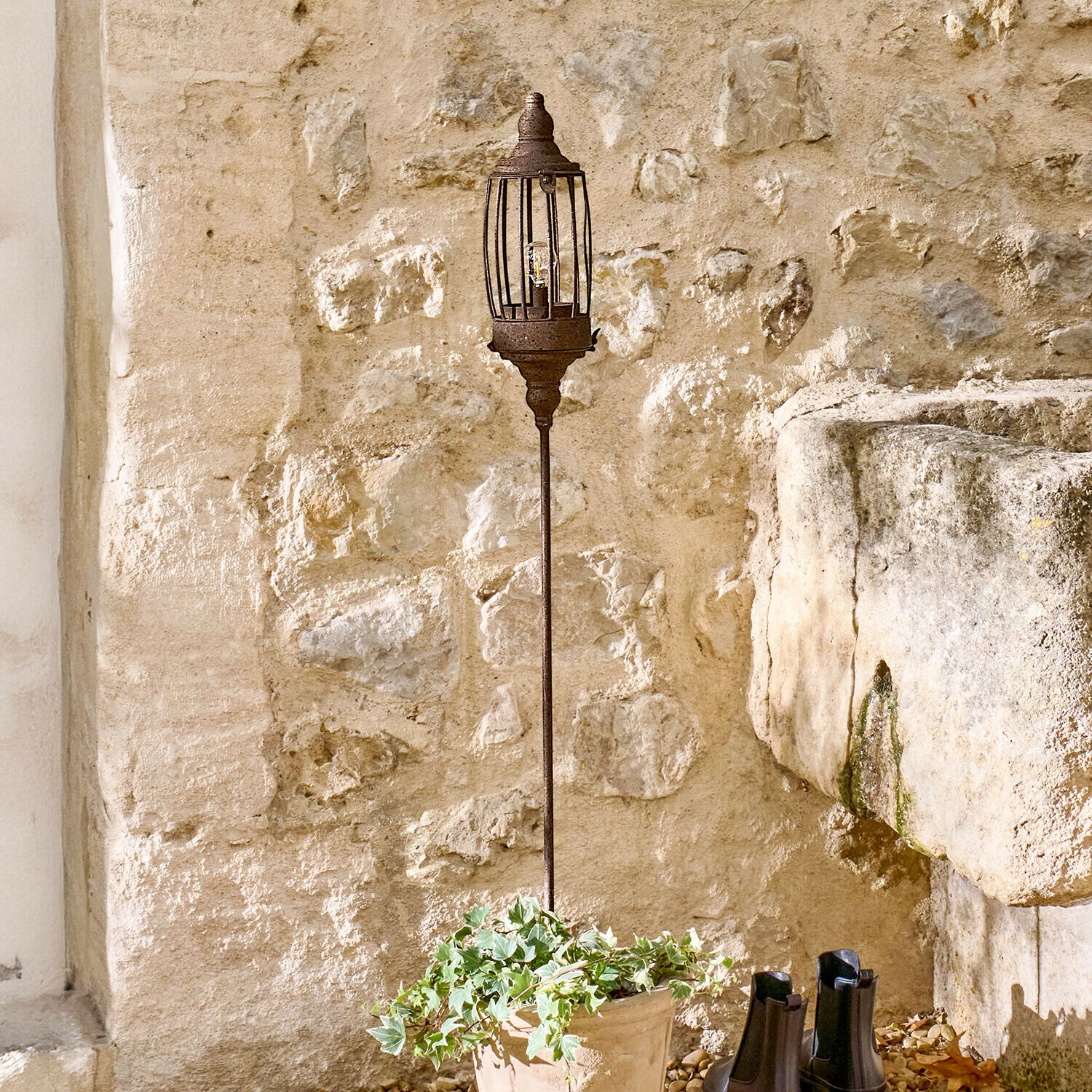 Lanterna a LED ricaricabile Lagia Lampada da giardino in metallo arrugginito su palo lungo davanti a muro in pietra rustica
