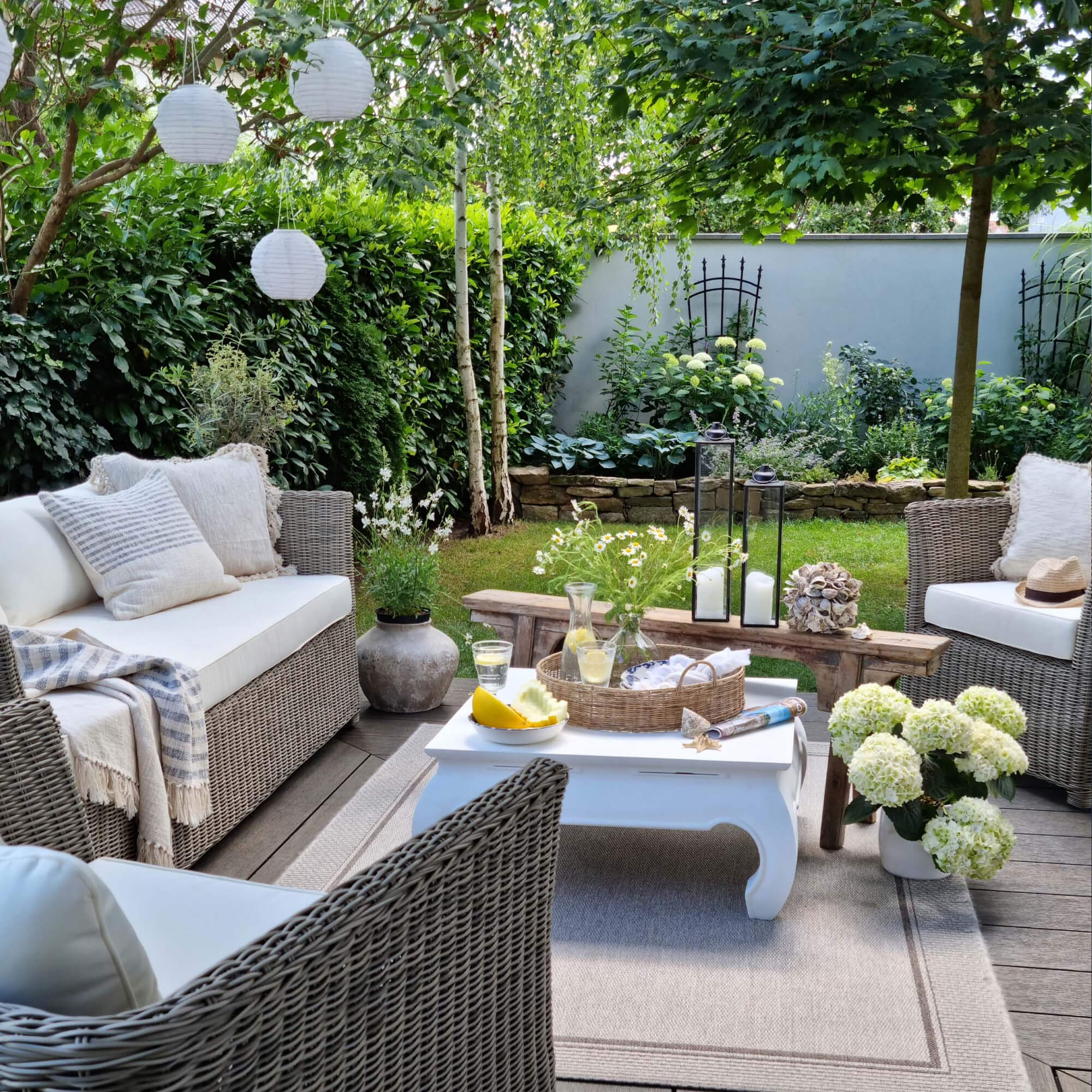 Angolo giardino accogliente con mobili in rattan grigio e cuscini bianchi, decorazioni e piante verdi.