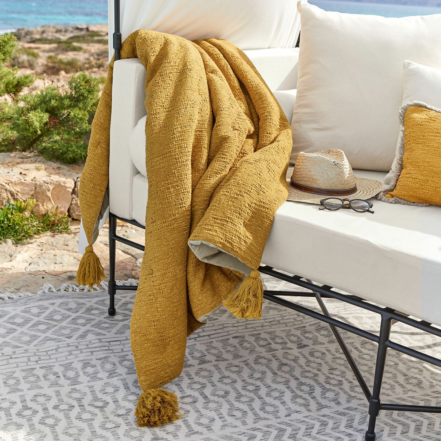 Trapuntino Rabea Plaid giallo con nappine su divano bianco da esterno, decorazione terrazza con occhiali e cappello