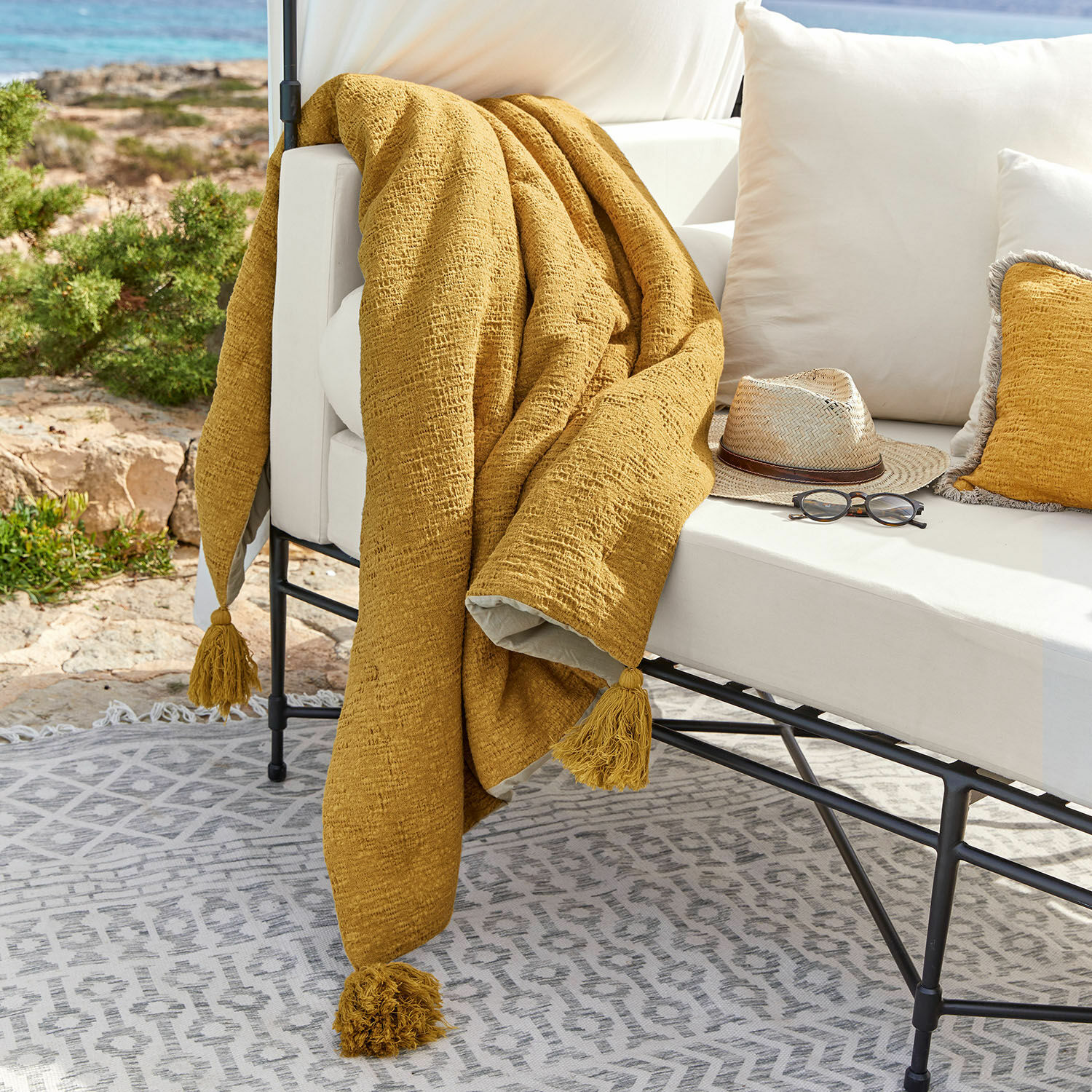 Trapuntino Rabea Plaid giallo con nappine su divano bianco da esterno, decorazione terrazza con occhiali e cappello