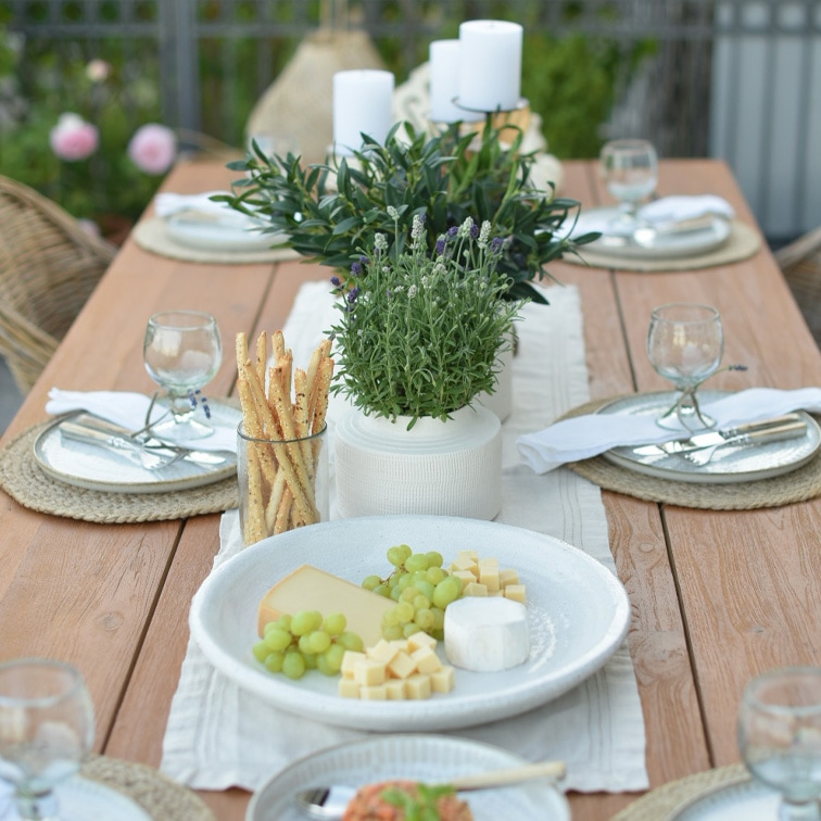 Tavolo in legno con stoviglie bianche, formaggi, uva verde e decorazione vegetale per pranzo all'aperto