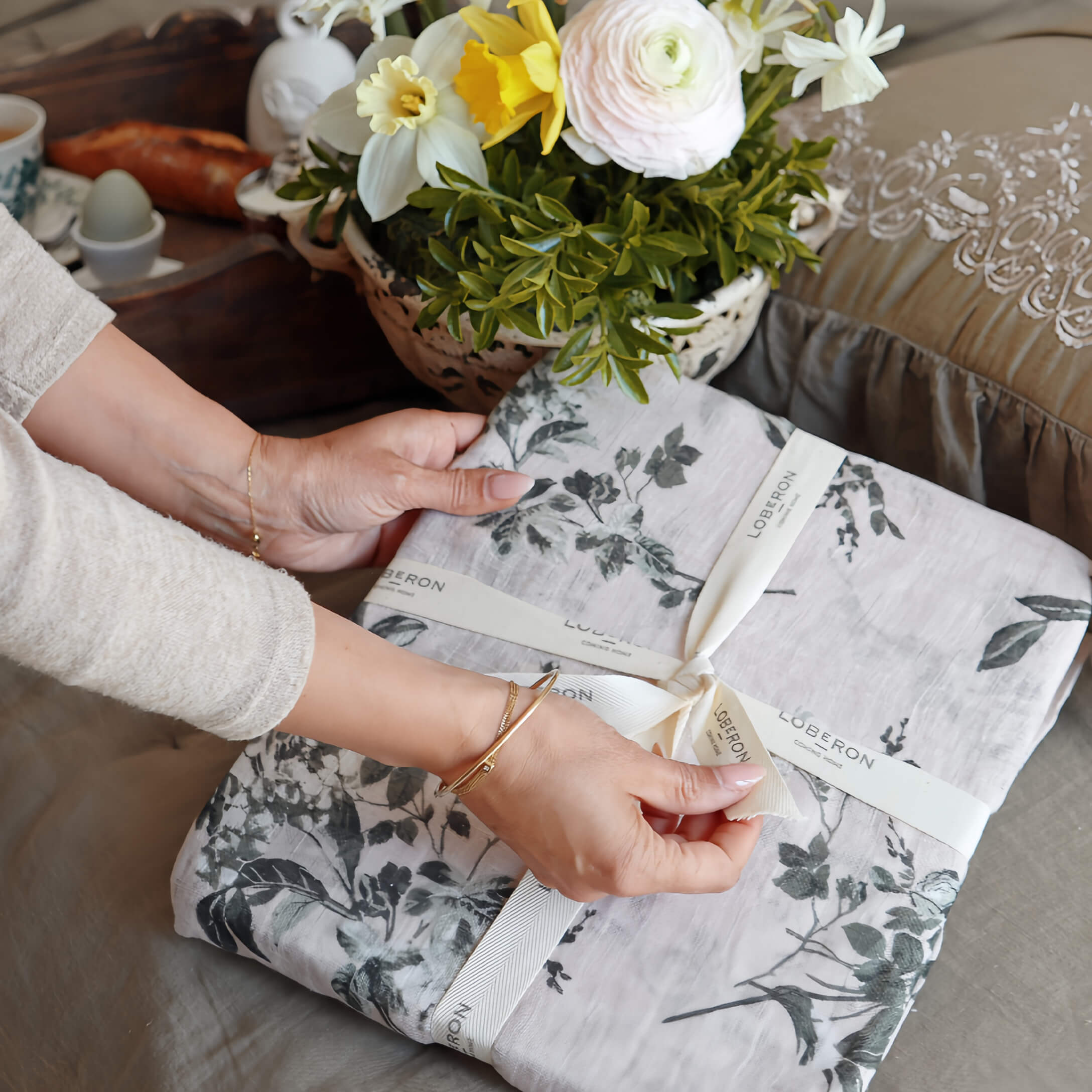 Mani che scartano biancheria da letto in lino grigio con motivo vegetale, fiori decorativi sul tavolo
