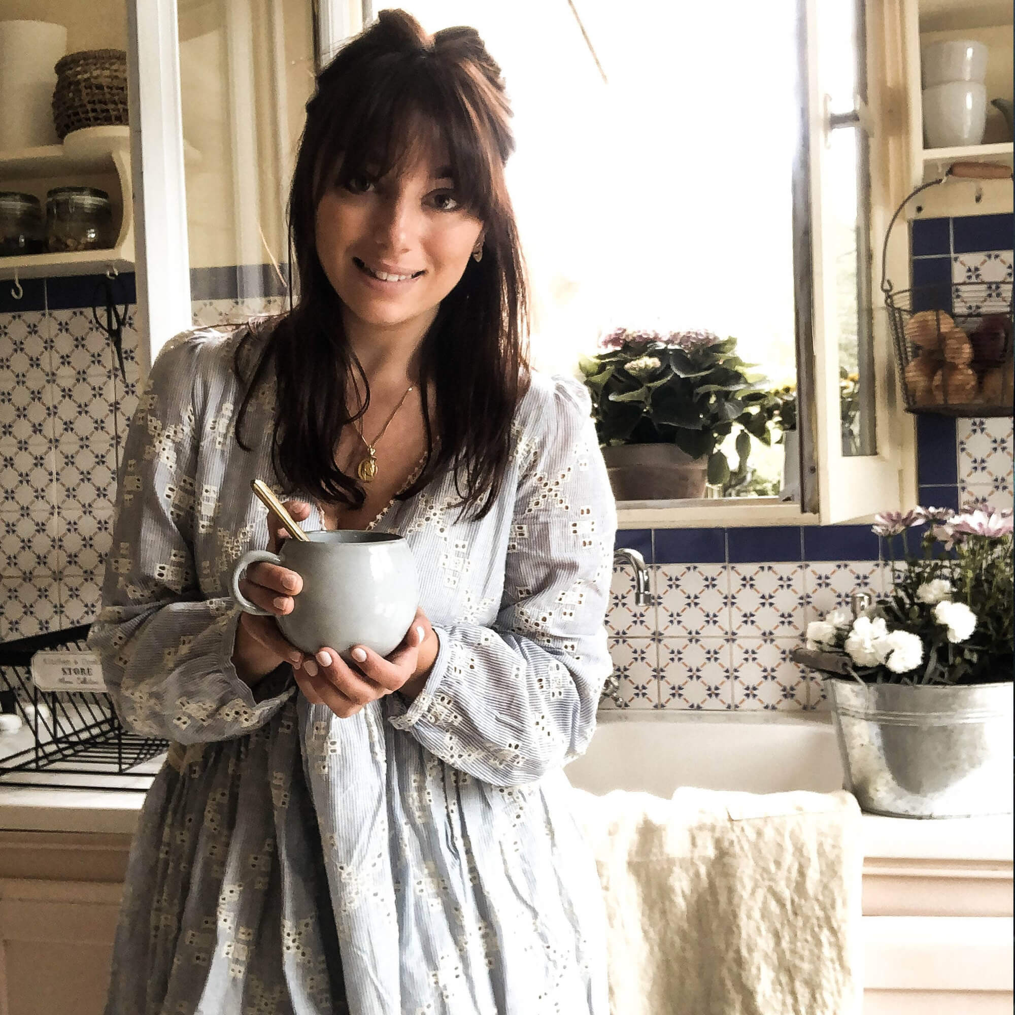 Donna in abito chiaro che tiene una tazza di ceramica in cucina con piastrelle blu e bianche e fiori