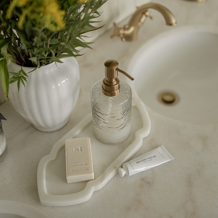 Dispenser di sapone in vetro con pompa in ottone su vassoio bianco accanto a sapone e crema mani su lavabo in marmo