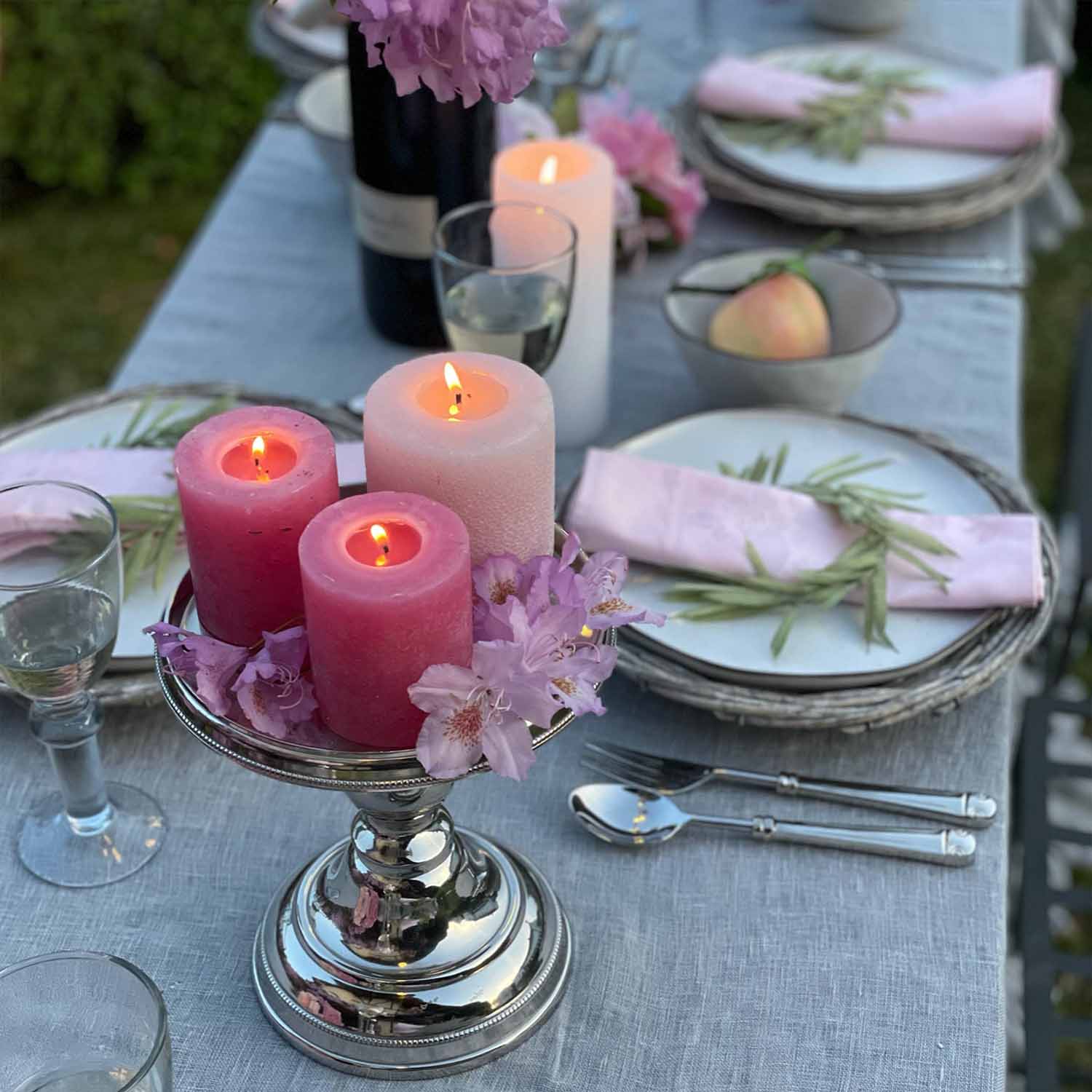 Portacandele argentato con tre candele accese rosa e bianca su tovaglia grigia con decorazioni floreali