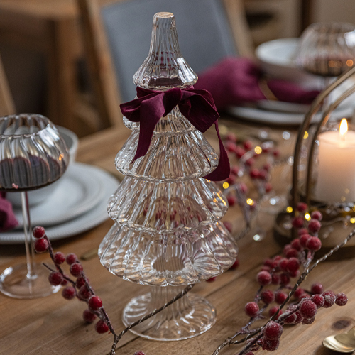 Decorazione natalizia a forma di albero in vetro con fiocco bordeaux su tavolo in legno con addobbi