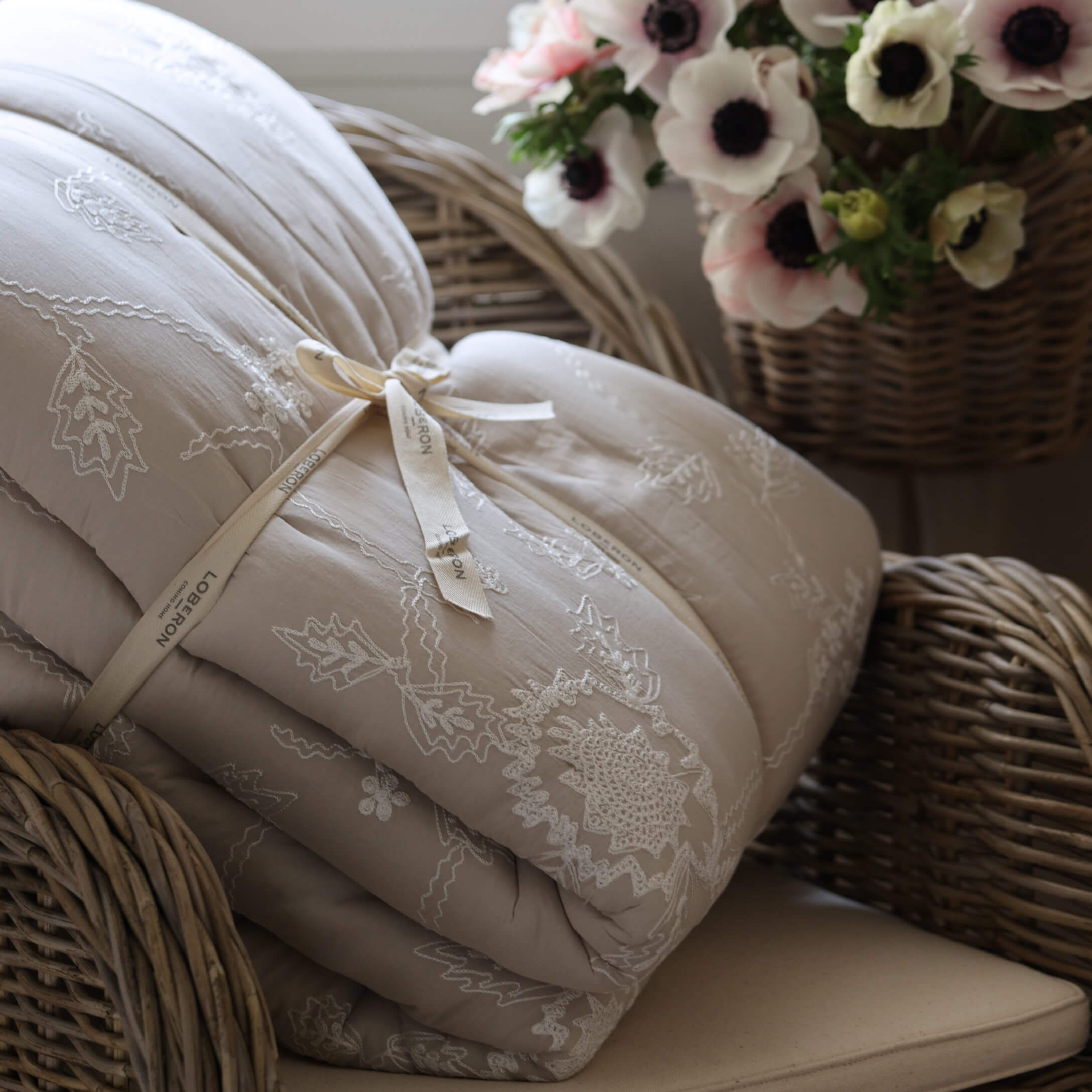 Trapunta beige trapuntata con ricamo su poltrona in vimini accanto a cesto di fiori, decorazione casa