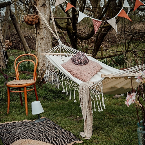 Amaca in tessuto bianco con cuscino rosa e coperta su prato verde, sedia in legno e ghirlanda decorativa