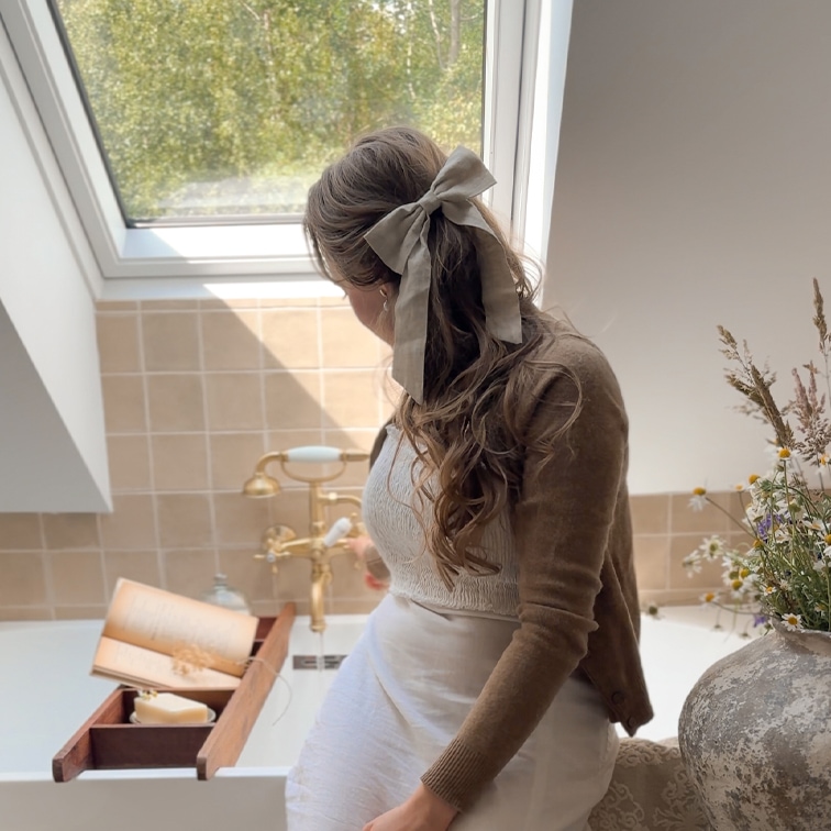 Donna con fiocco di lino beige nei capelli seduta vicino a vasca con vassoio in legno e libro, luce naturale