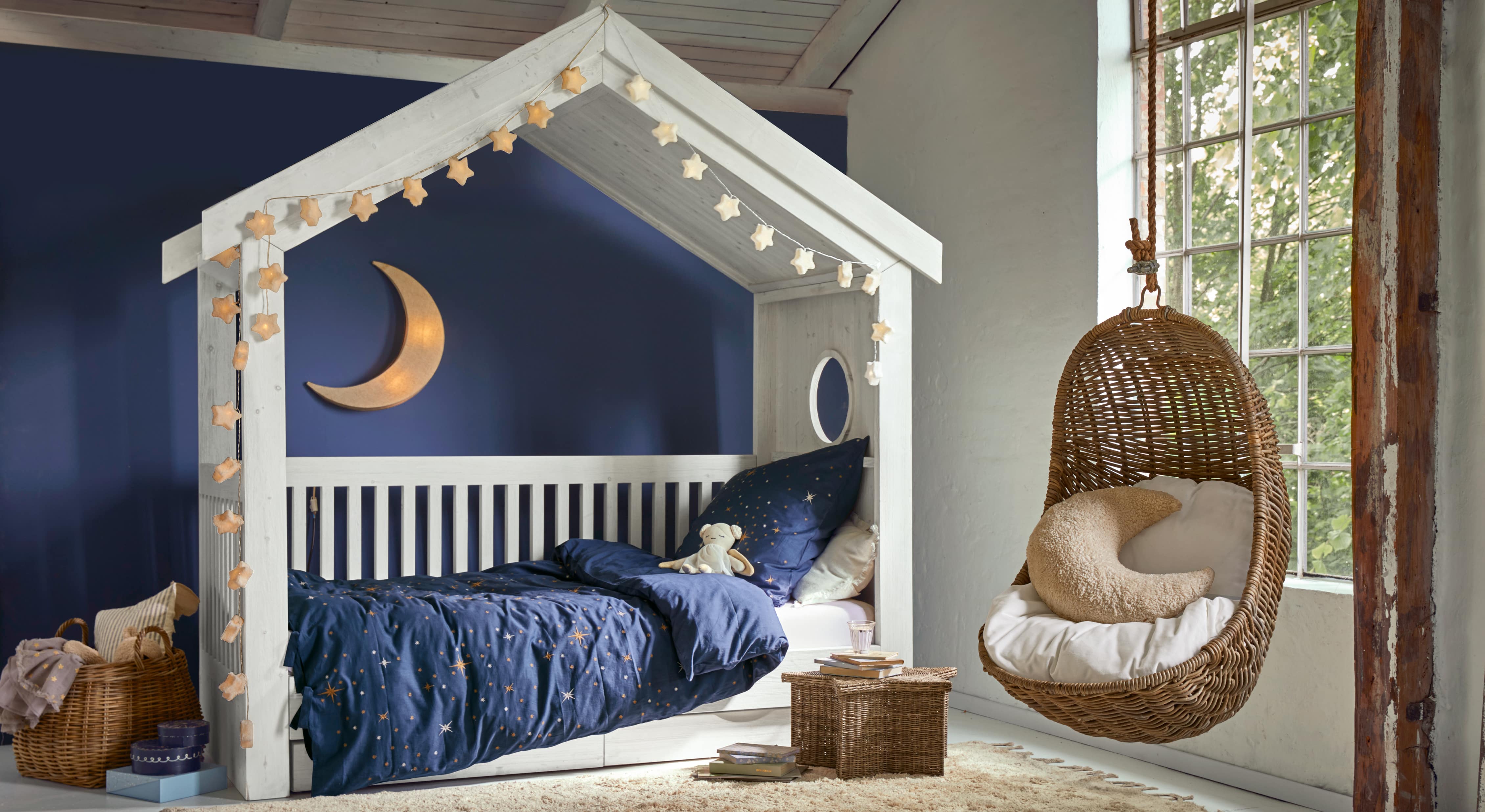 Letto in legno bianco a forma di casetta con biancheria blu stellata e poltrona sospesa in vimini