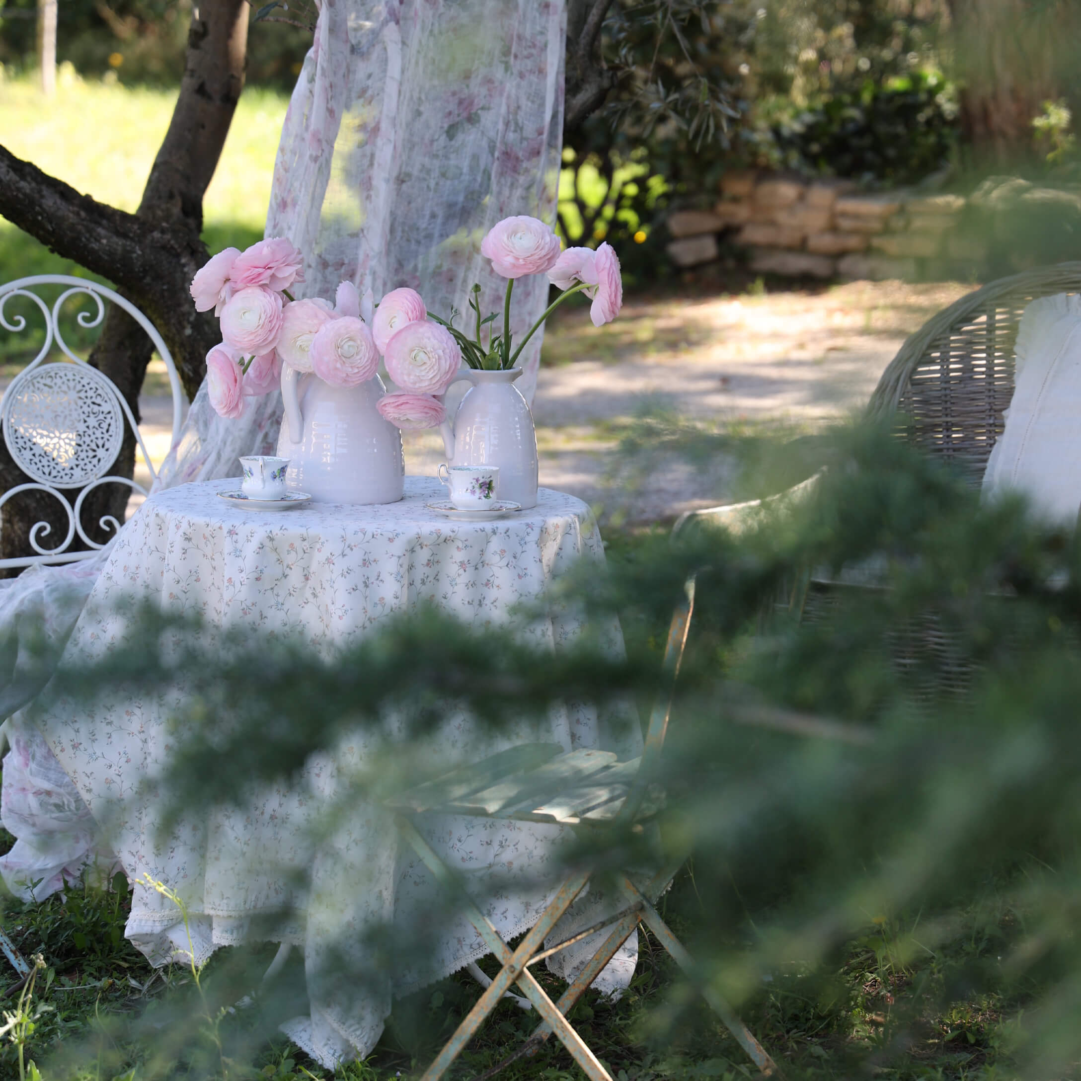 Tavolo da giardino bianco con tovaglia floreale e brocche bianche con fiori rosa, atmosfera romantica