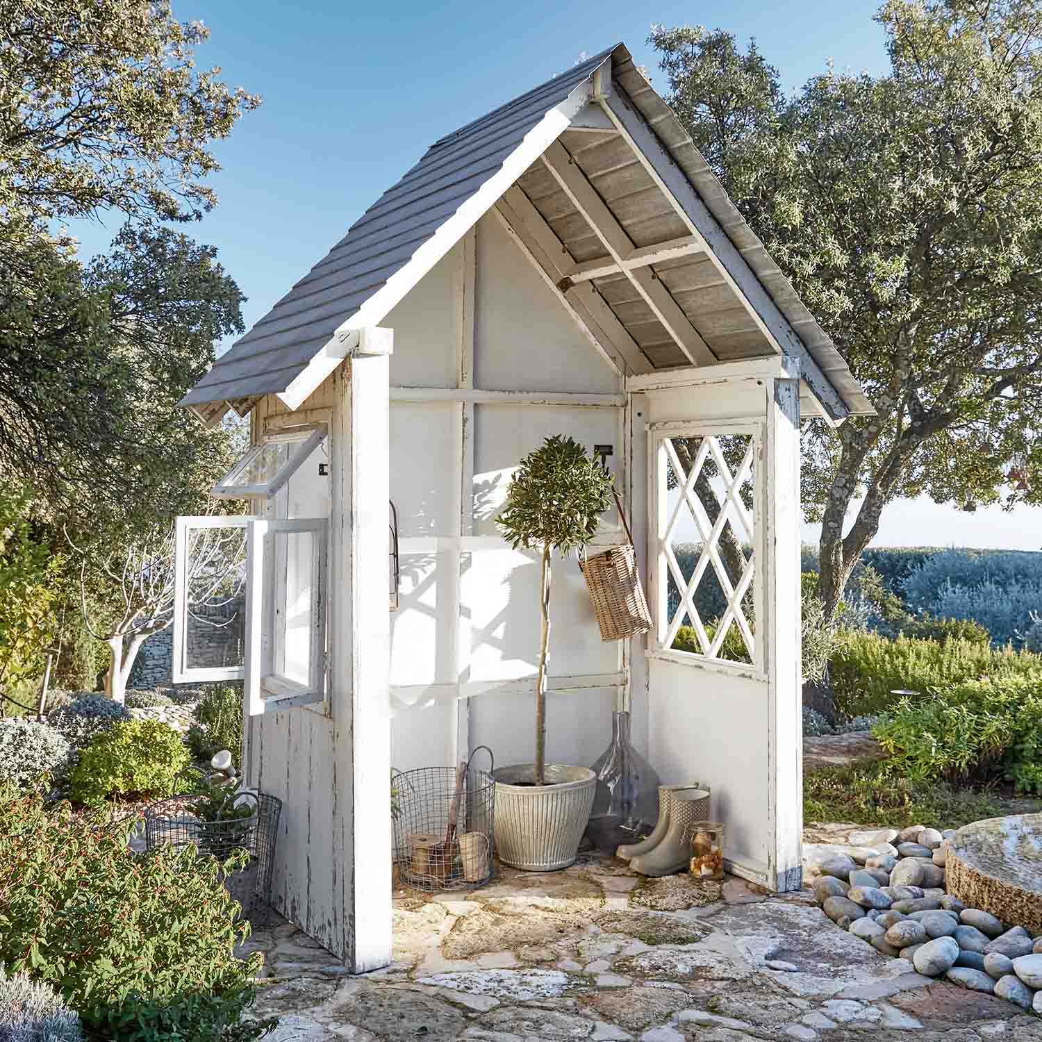 Piccola casetta da giardino in legno bianco con finestre e piante su pavimento in pietra in giardino soleggiato