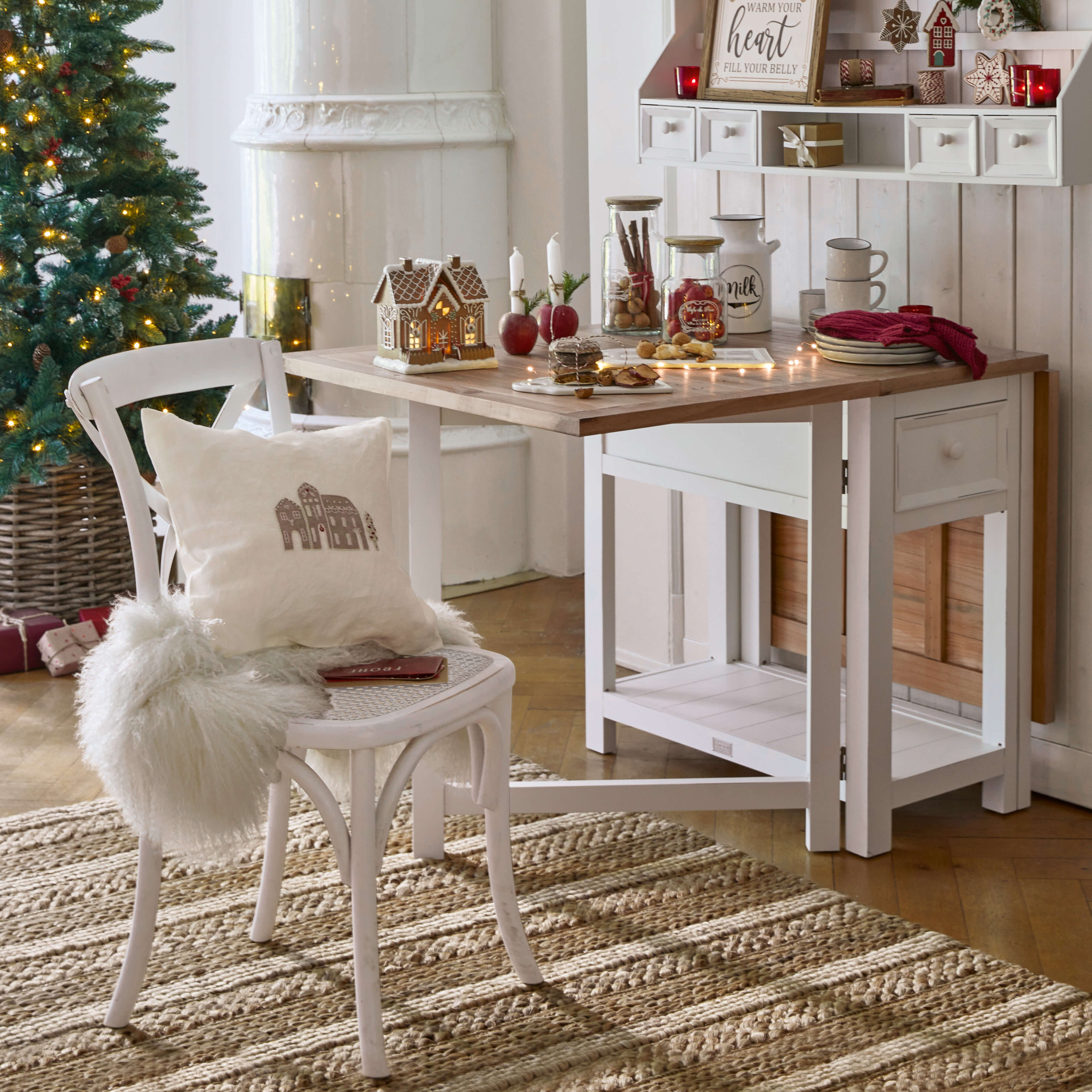 Tavolo e sedia in legno bianco con decorazioni natalizie e albero di Natale illuminato in ambiente accogliente