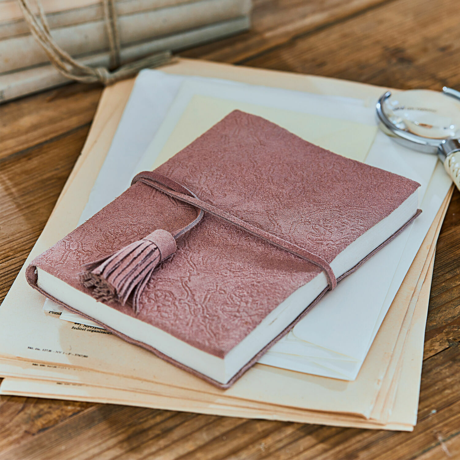 Taccuino Orcival Taccuino rosa in camoscio con nappina su tavolo in legno con documenti, accessori per scrivere