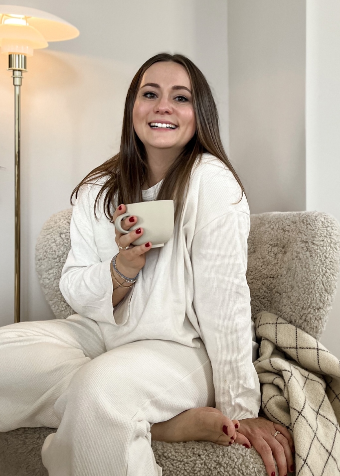 Donna con abbigliamento bianco morbido seduta su poltrona con coperta, tiene una tazza beige.