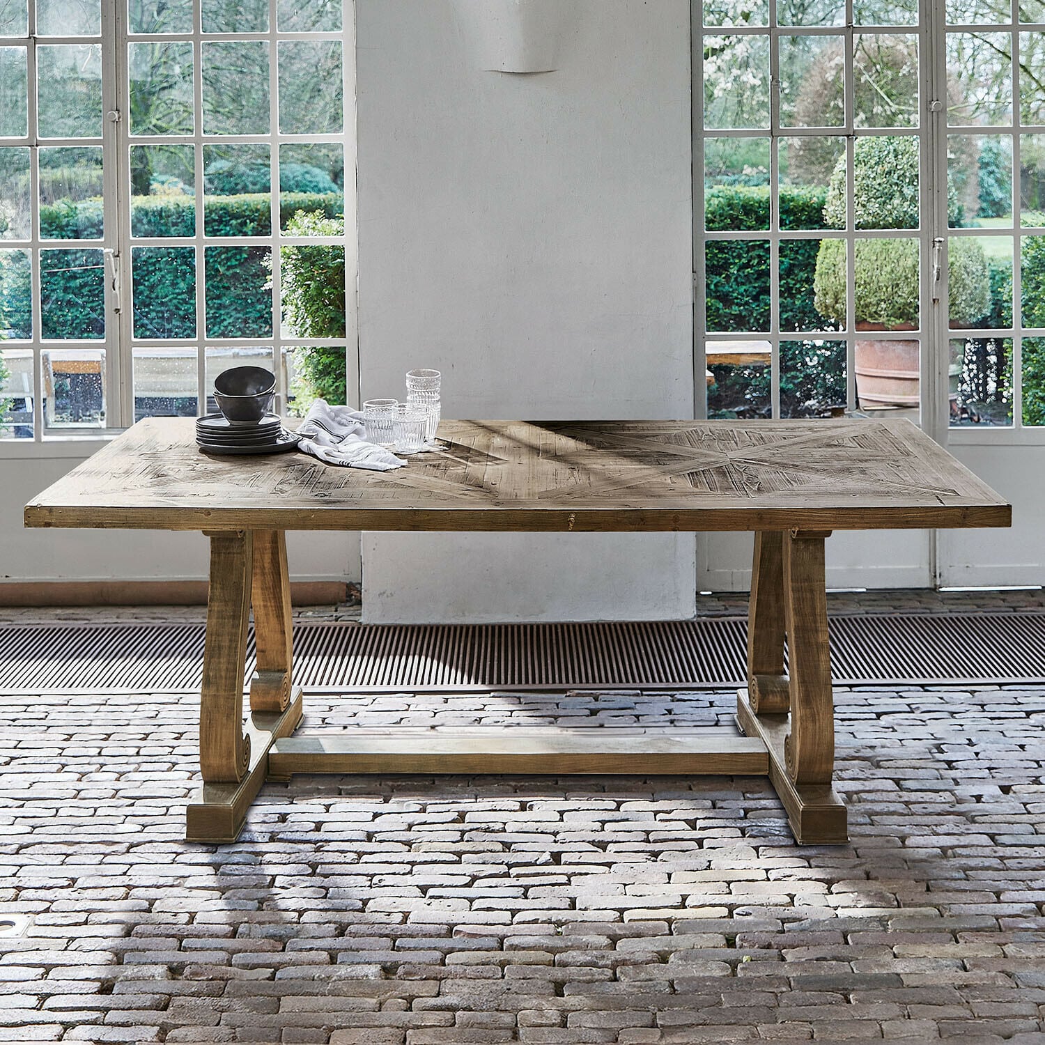 Tavolo Campbelltown Tavolo in legno colore naturale con piano decorativo davanti a grandi finestre e pavimento in mattoni