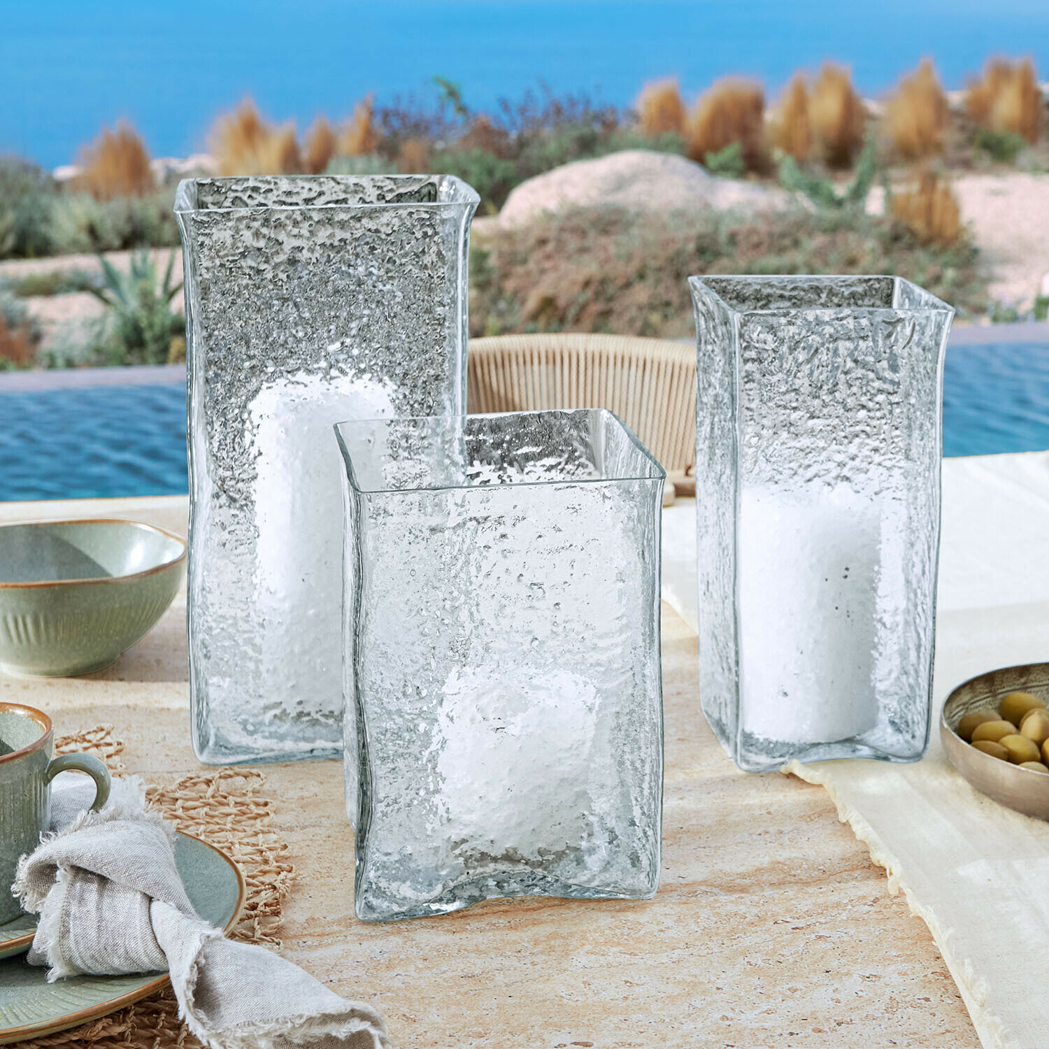 Set di 3 portacandele Xarlac Tre lanterne in vetro trasparente con texture e candele bianche su tavolo esterno vicino alla piscina