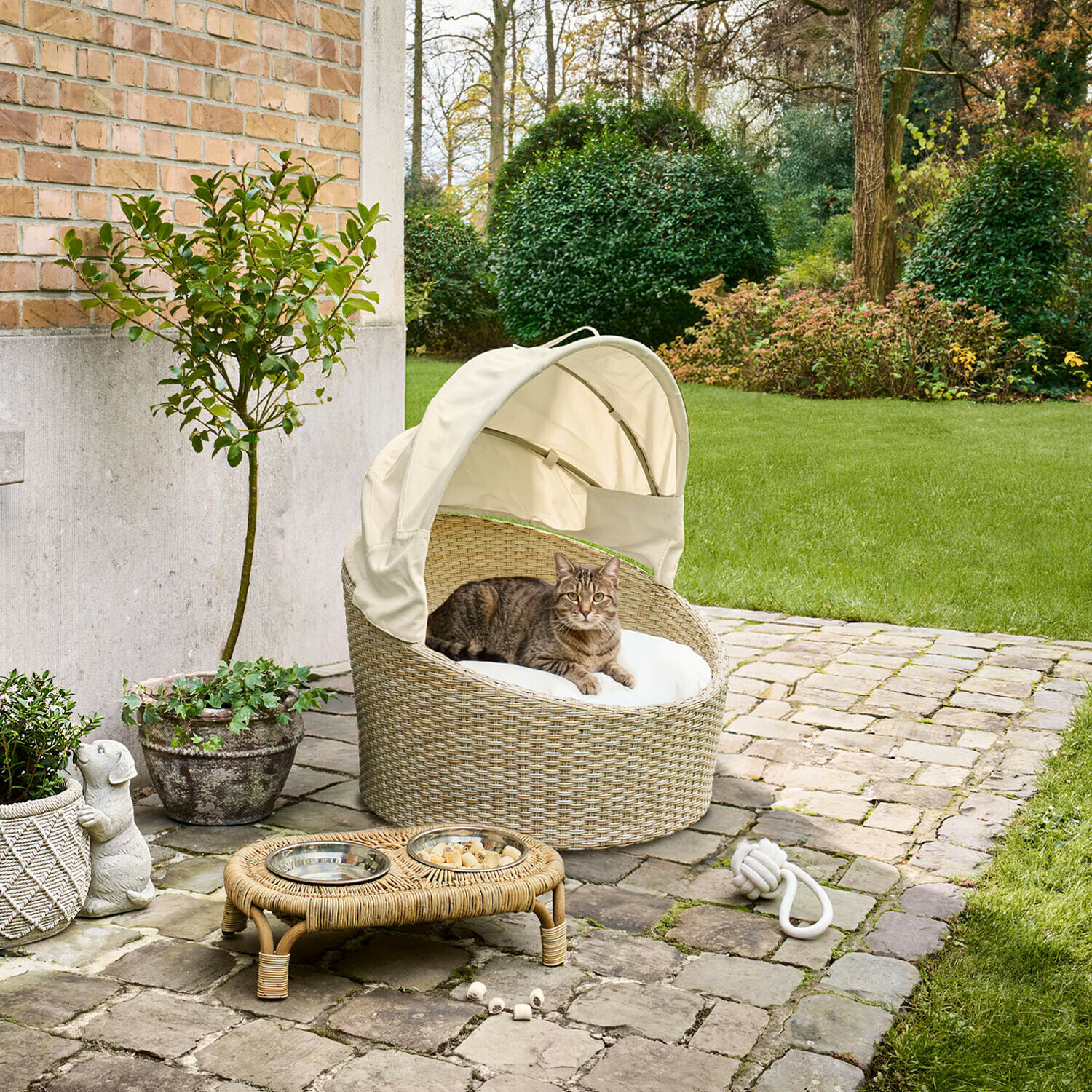 Cuccia per animali per interni ed esterni Brinelza Cuccia ovale per gatti in rattan beige con cuscino bianco e tettuccio, posizionata su terrazza in pietra