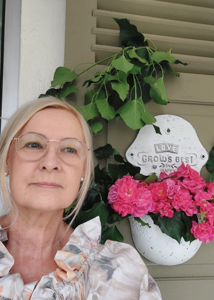 Donna con occhiali accanto a vaso bianco con fiori rosa e foglie verdi su muro