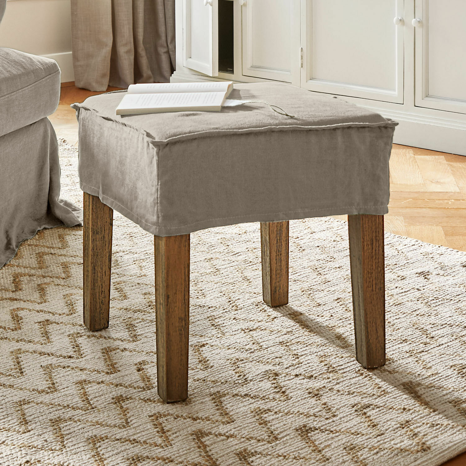 Sgabello Vionnique Pouf beige con rivestimento in cotone e gambe in legno su tappeto chiaro in soggiorno