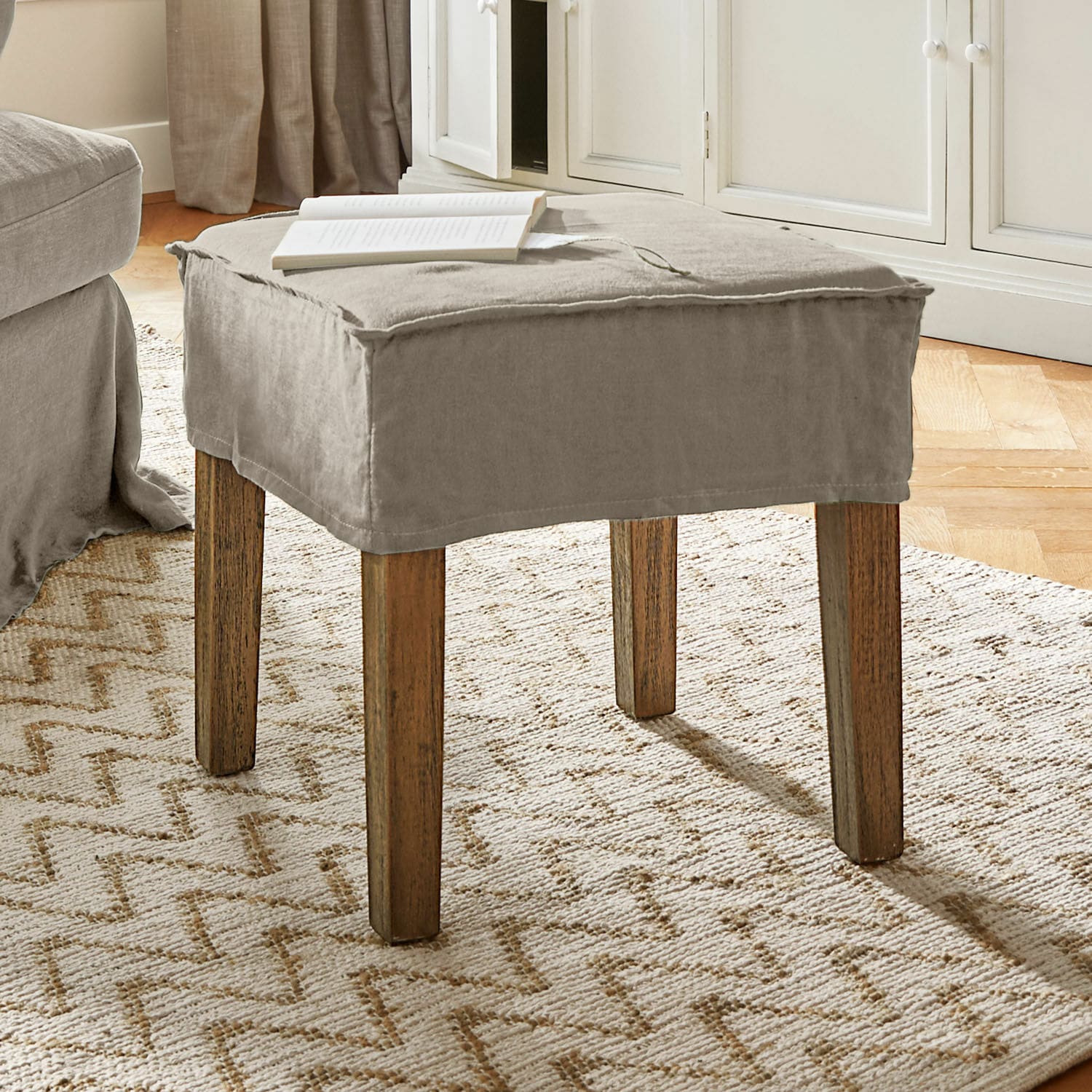Sgabello Vionnique Pouf beige con rivestimento in cotone e gambe in legno su tappeto chiaro in soggiorno