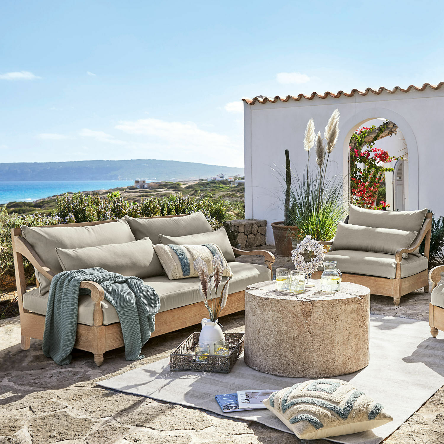 Salottino Montcassin Set di mobili da giardino in legno chiaro con cuscini beige su terrazza in pietra con vista mare