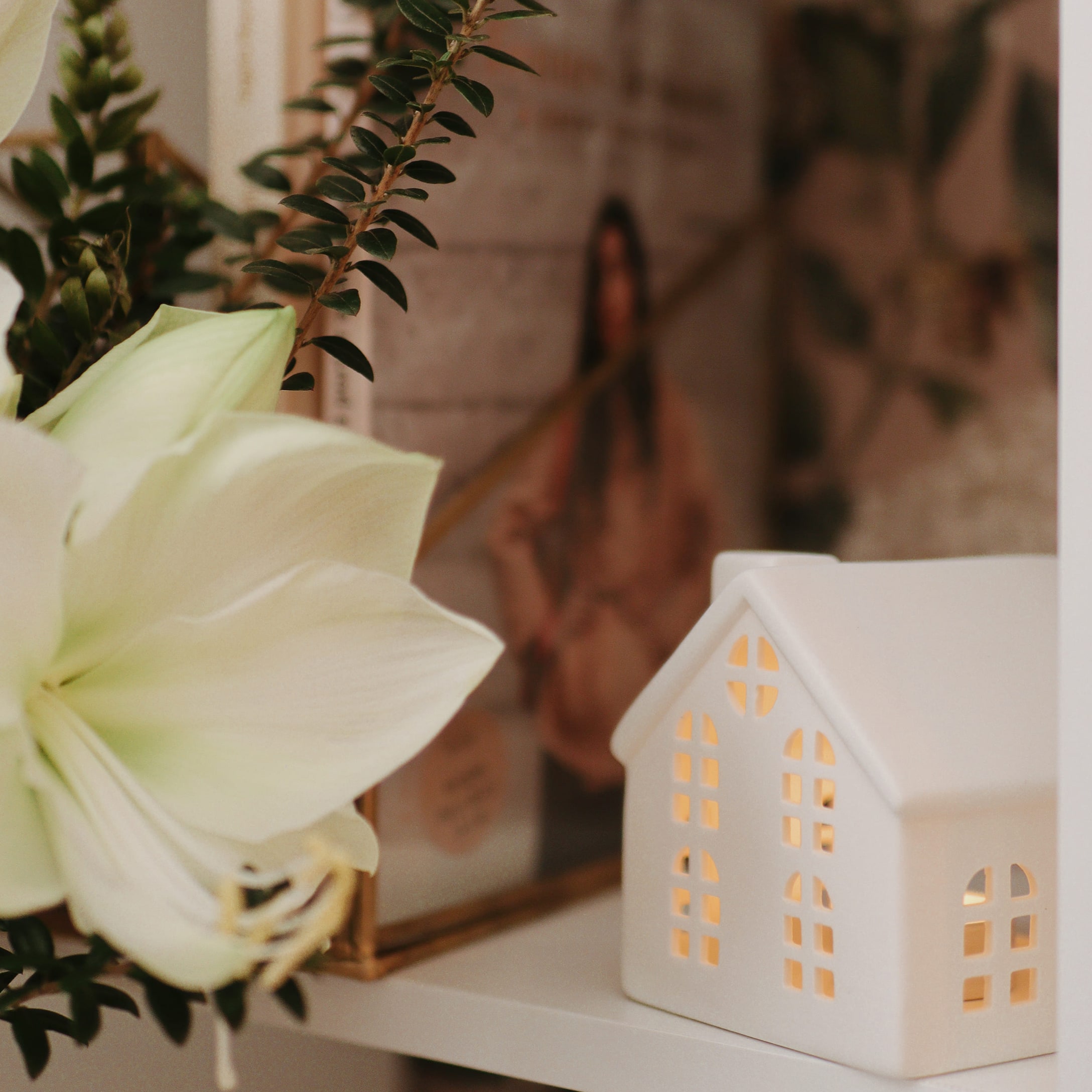 Decorazione casa in ceramica bianca illuminata su mensola accanto a fiore bianco e foglie verdi