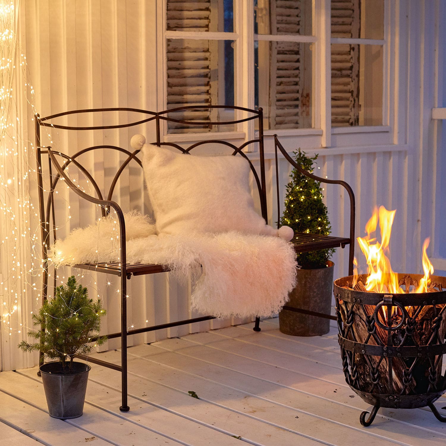 Panchina Jablines Panchina in metallo con cuscino bianco e coperta di pelliccia accanto a un braciere su terrazza con luci decorative