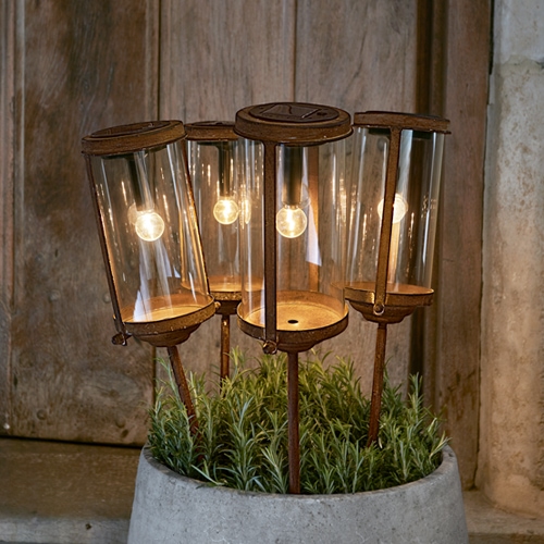 Set di quattro lanterne da giardino in vetro e metallo arrugginito in vaso con piante verdi, illuminazione esterna decorativa