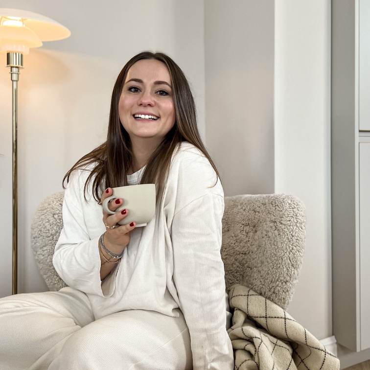 Donna sorridente con abbigliamento bianco morbido seduta su poltrona con tazza in ambiente accogliente