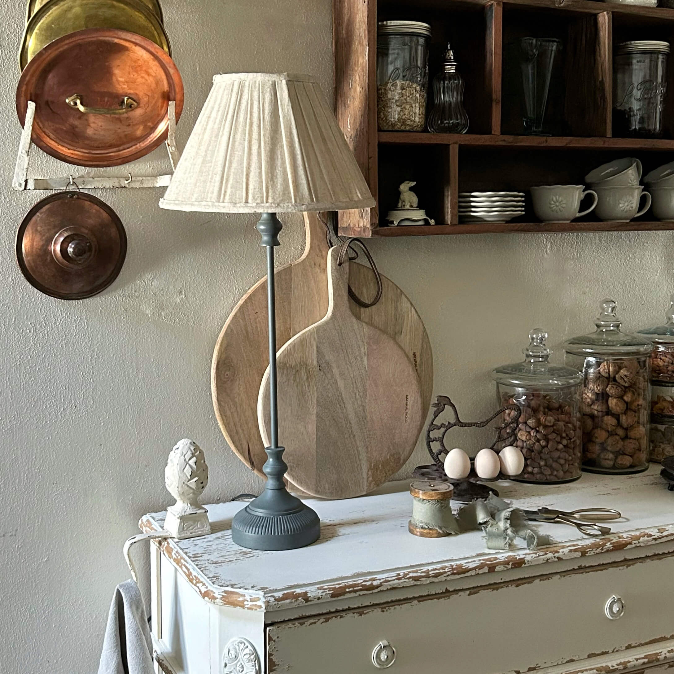 Lampada da tavolo con base grigia e paralume crema su comò bianco invecchiato in cucina rustica