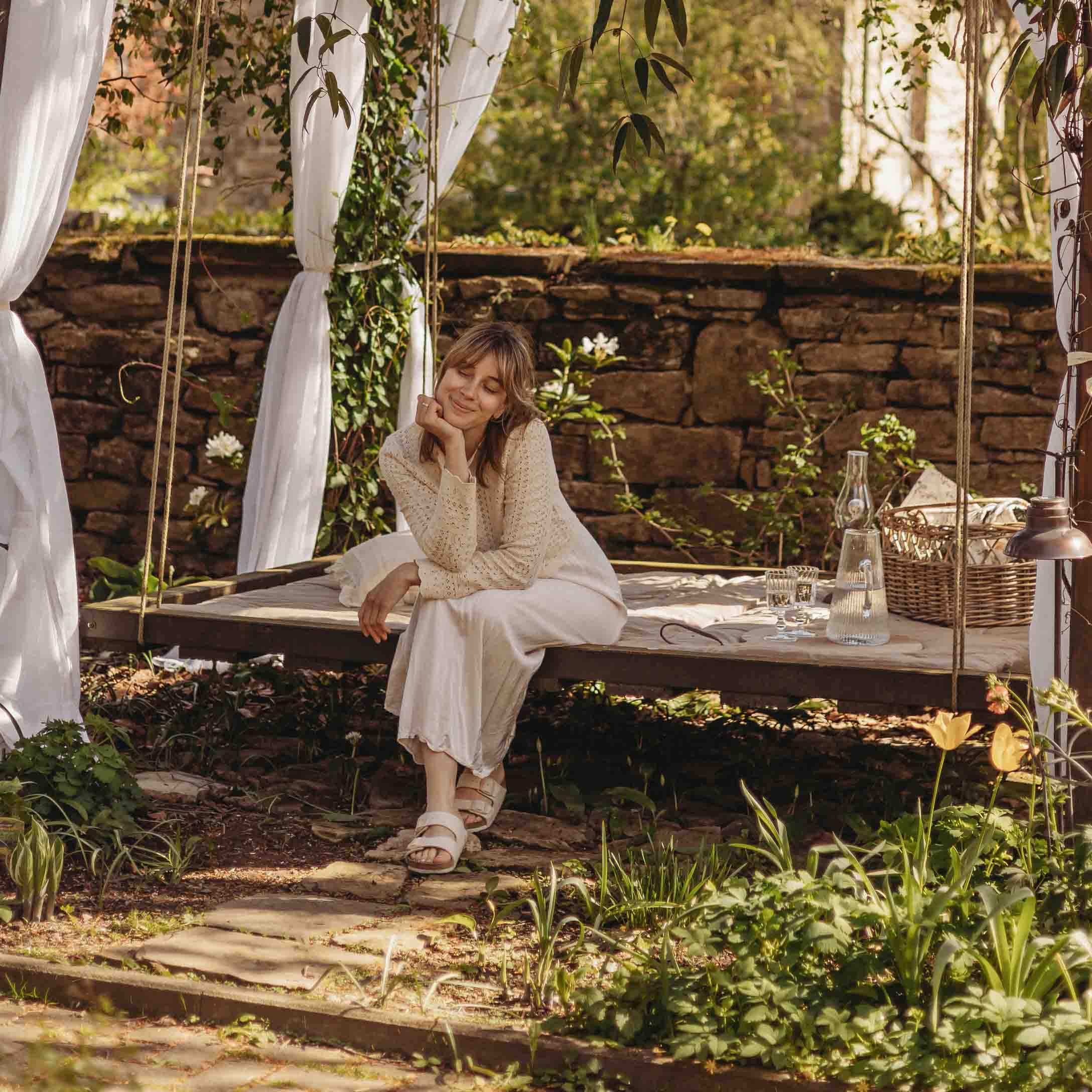 Donna con maglione beige seduta su un'altalena da giardino in legno con tende bianche e ambiente naturale