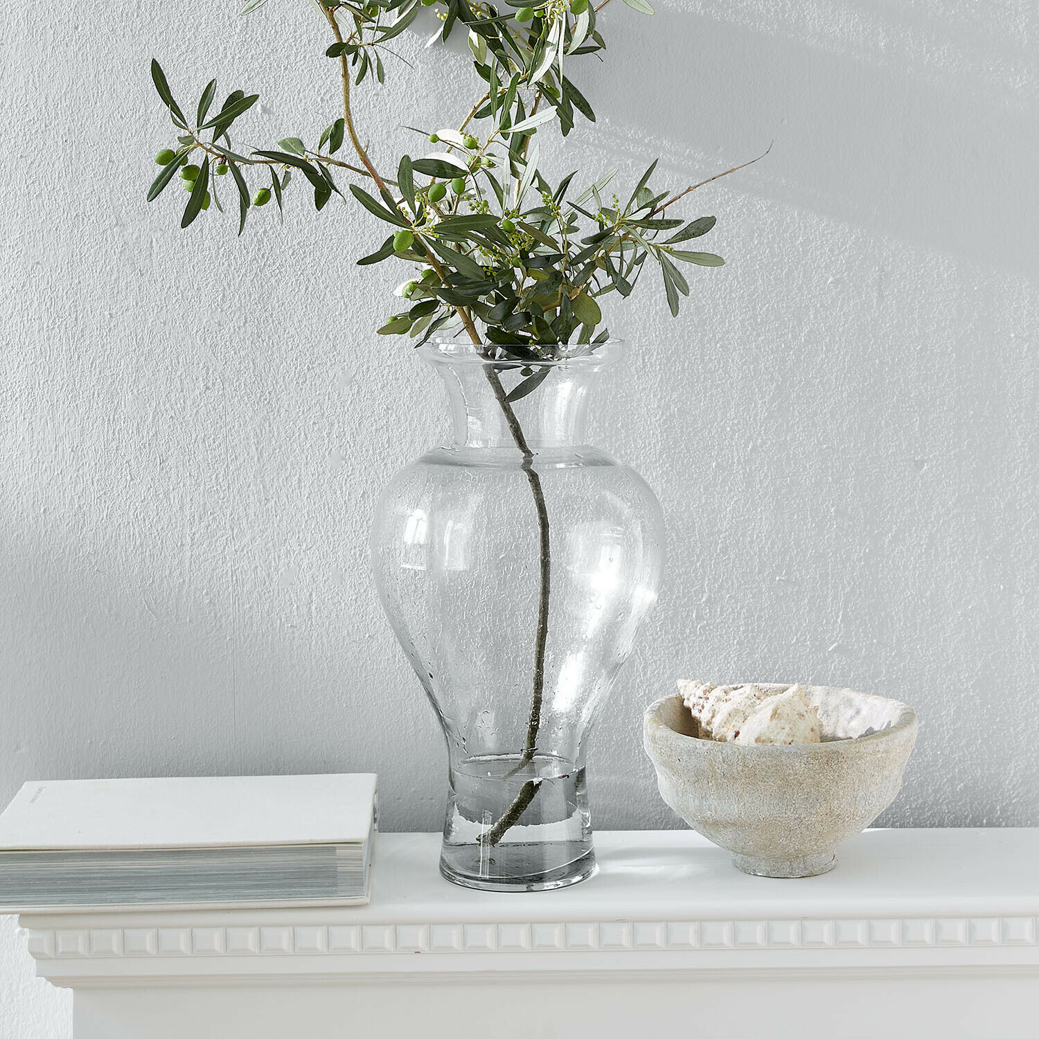 Vaso in vetro trasparente con rami verdi su mensola bianca con libro e ciotola con conchiglie