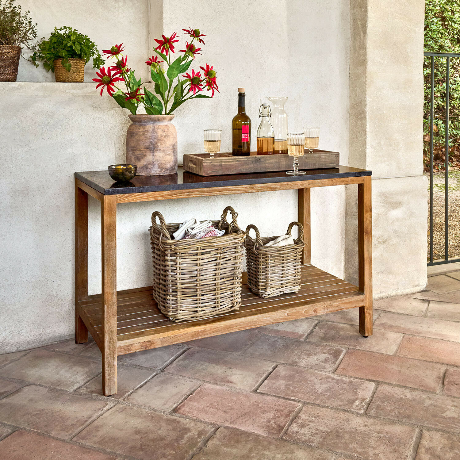Tavolo da cucina per esterni Corviano Consolle in legno con piano nero, decorata con vaso di fiori rossi e cesti di vimini sul ripiano inferiore