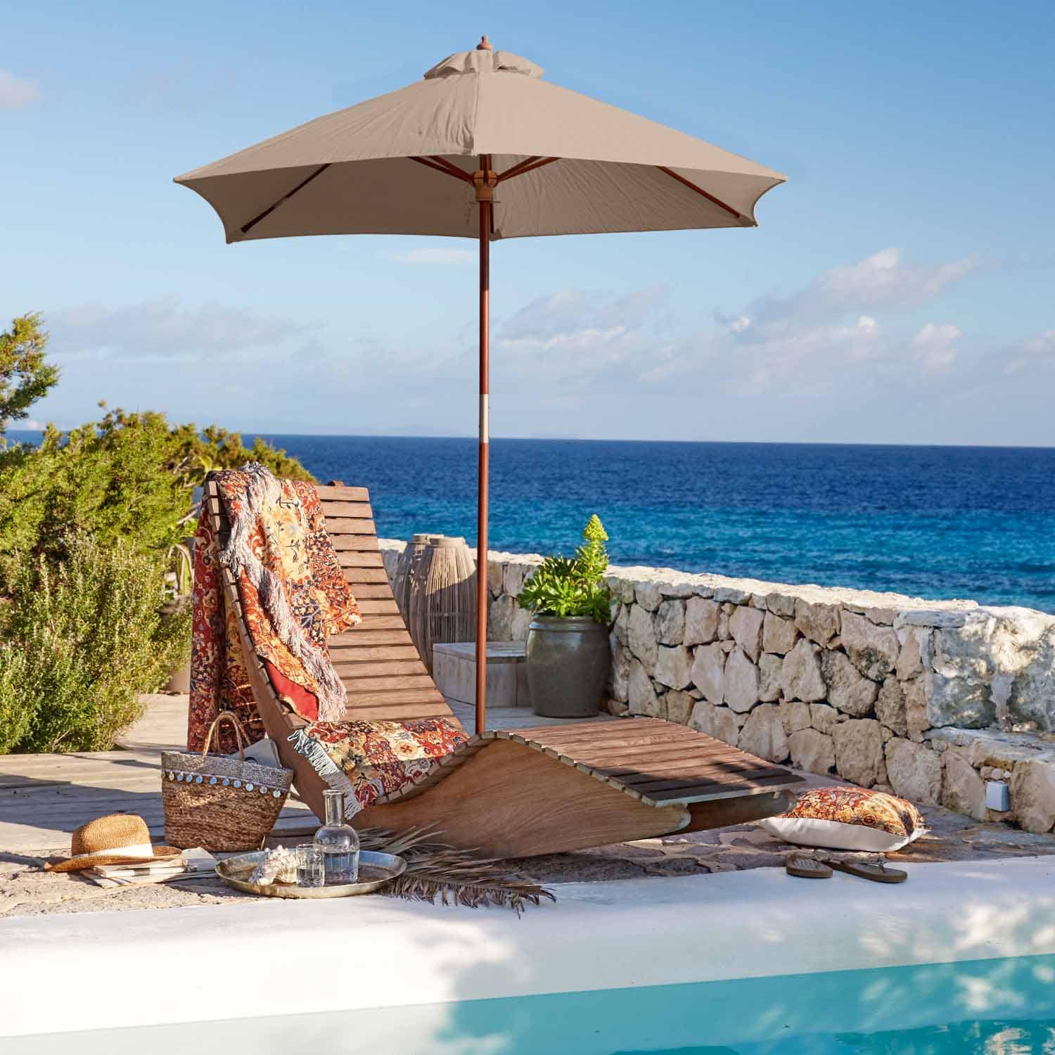 Lettino in legno con ombrellone beige e coperta colorata vicino alla piscina con vista mare