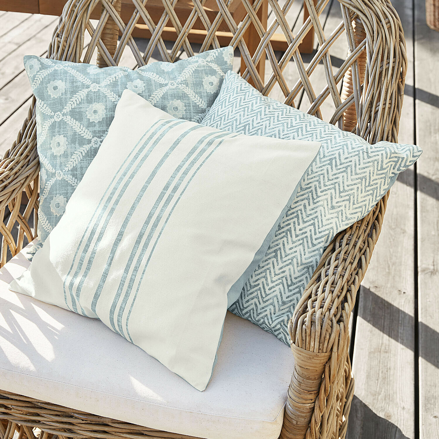 Tre cuscini decorativi in tonalità di azzurro e bianco su una poltrona in vimini su terrazza in legno