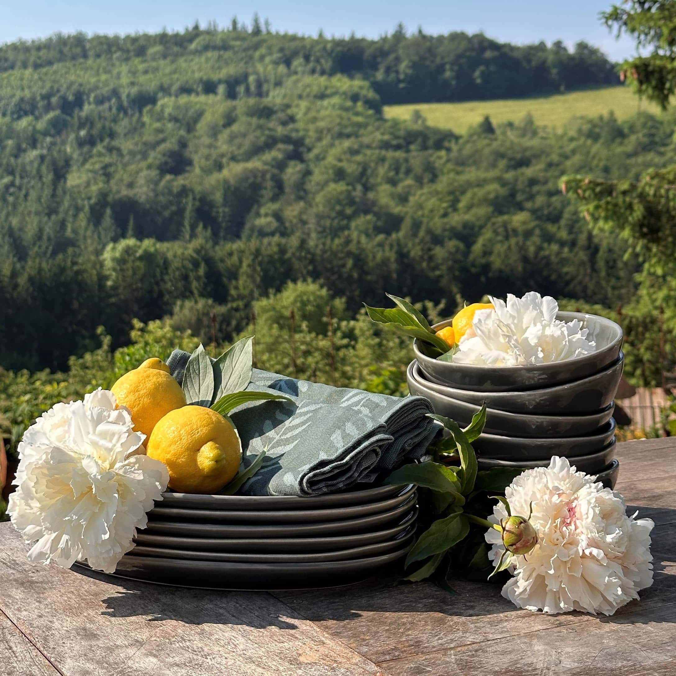 Set di piatti e ciotole in ceramica grigia con fiori bianchi e limoni su tavolo di legno davanti a foresta verde