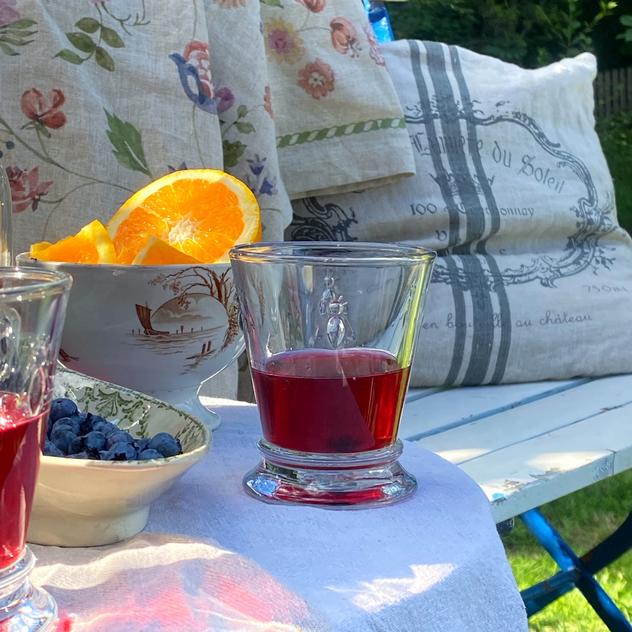 Bicchiere con bevanda rossa su tovaglia bianca, accanto ciotola con mirtilli e arancia su tavolo da giardino