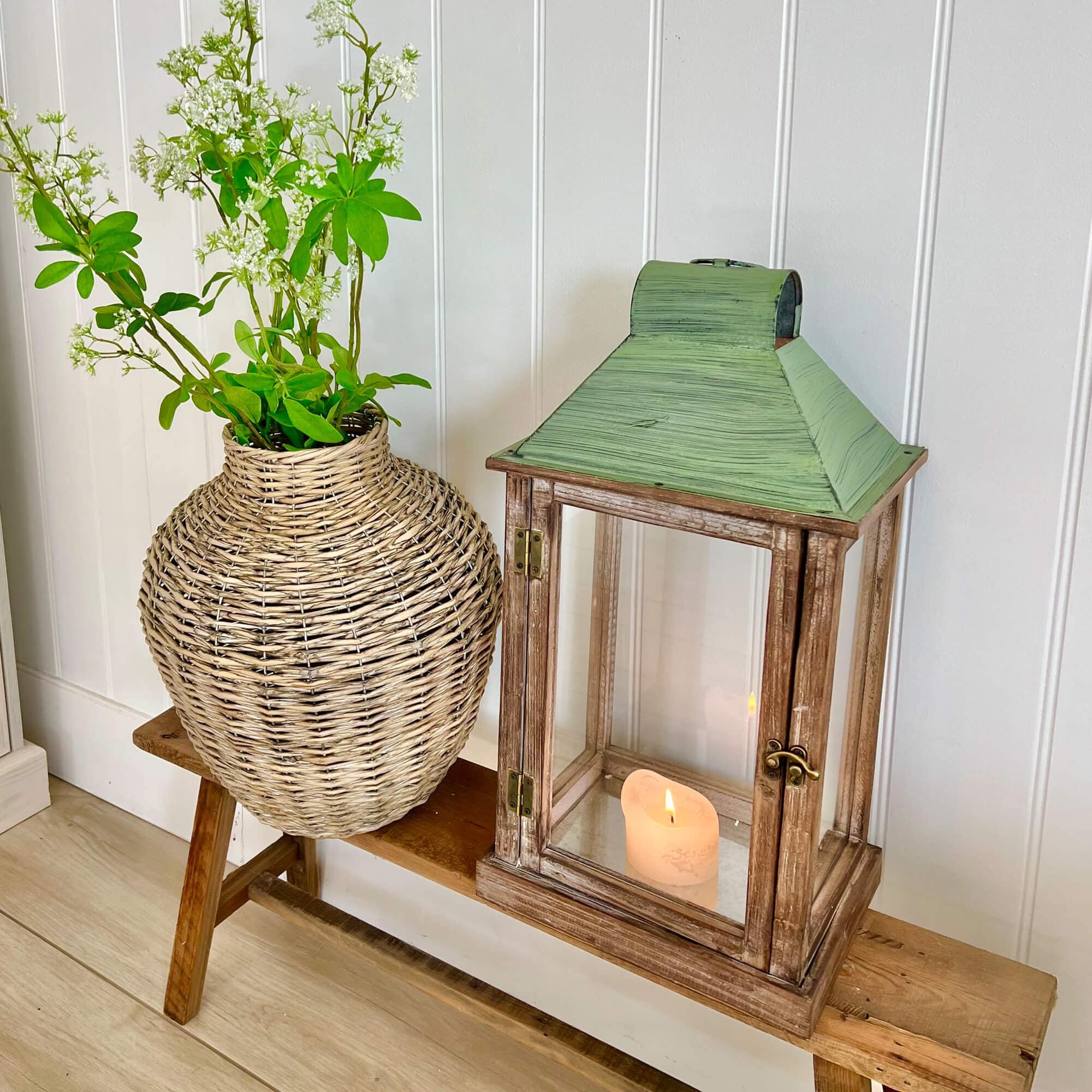 Lanterna rustica in legno con tetto in metallo verde e vaso in vimini con fiori bianchi su panca in legno