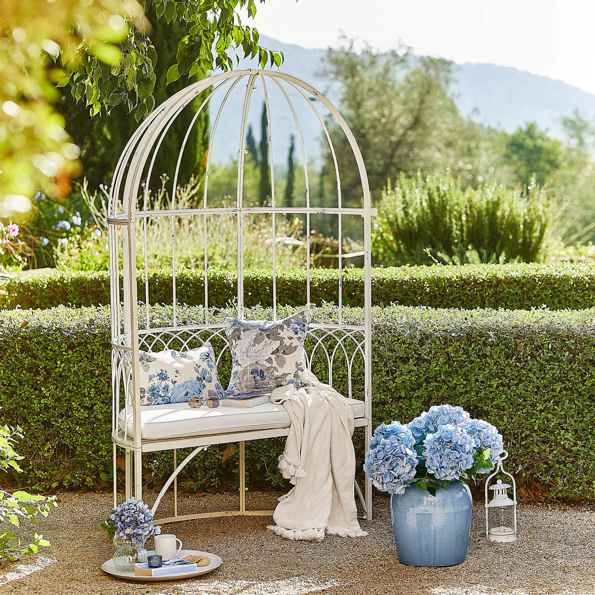 Panca da giardino in metallo bianco con cuscini e fiori blu in vaso su sfondo verde