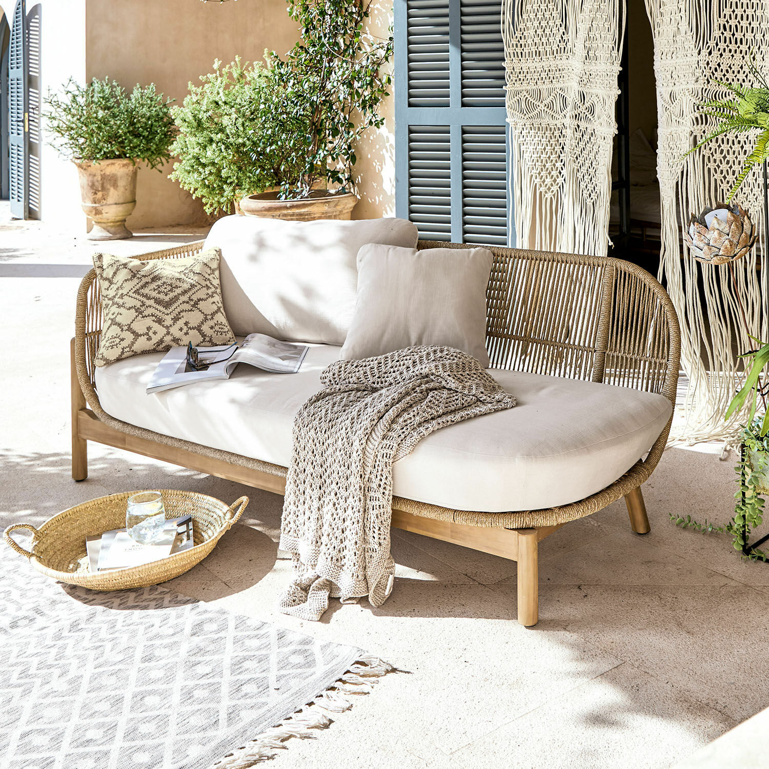 Chaise-longue FSC®-100% Belvidere Divano in rattan e legno con cuscini crema e coperta su terrazza luminosa con piante