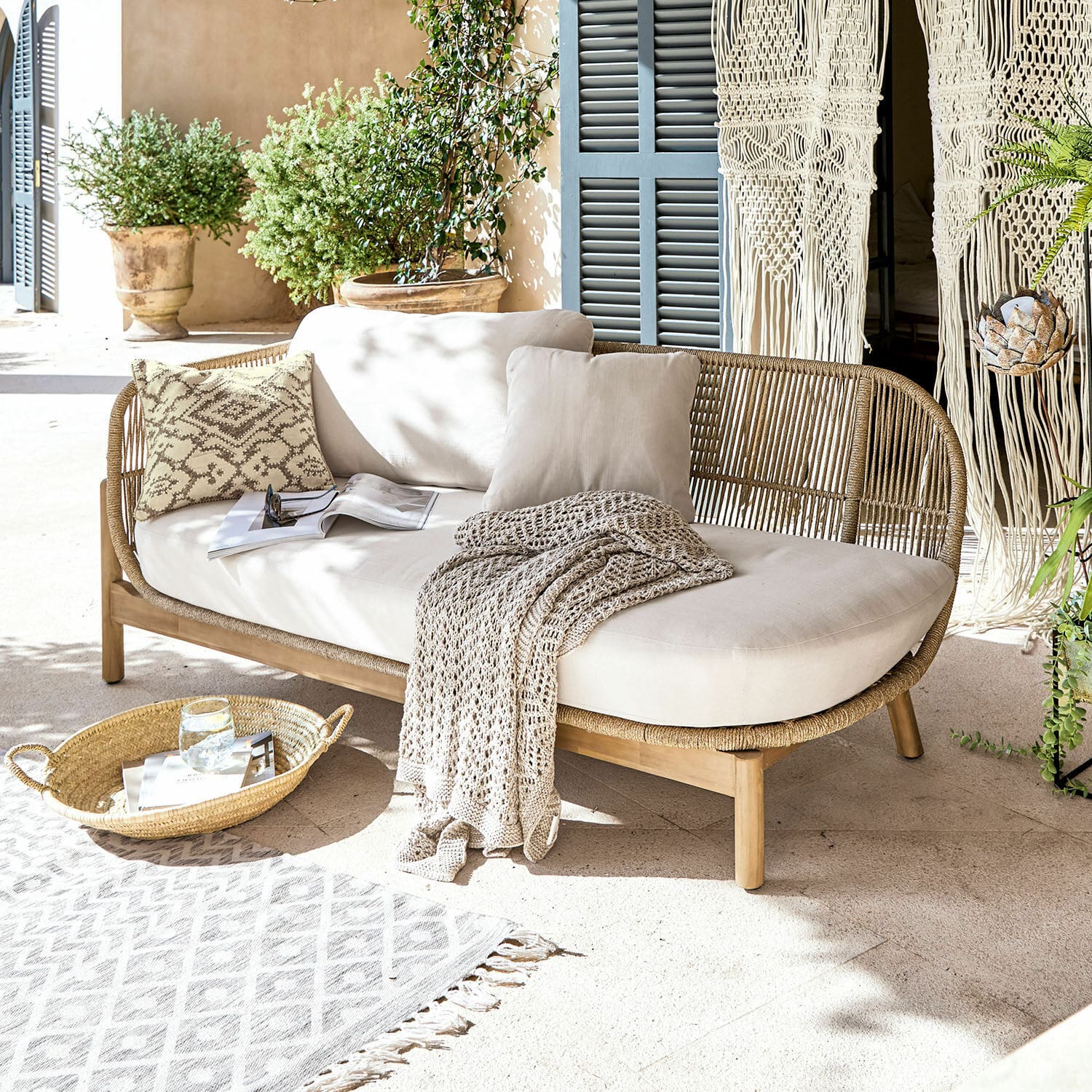 Chaise-longue FSC®-100% Belvidere Divano in rattan e legno con cuscini crema e coperta su terrazza luminosa con piante