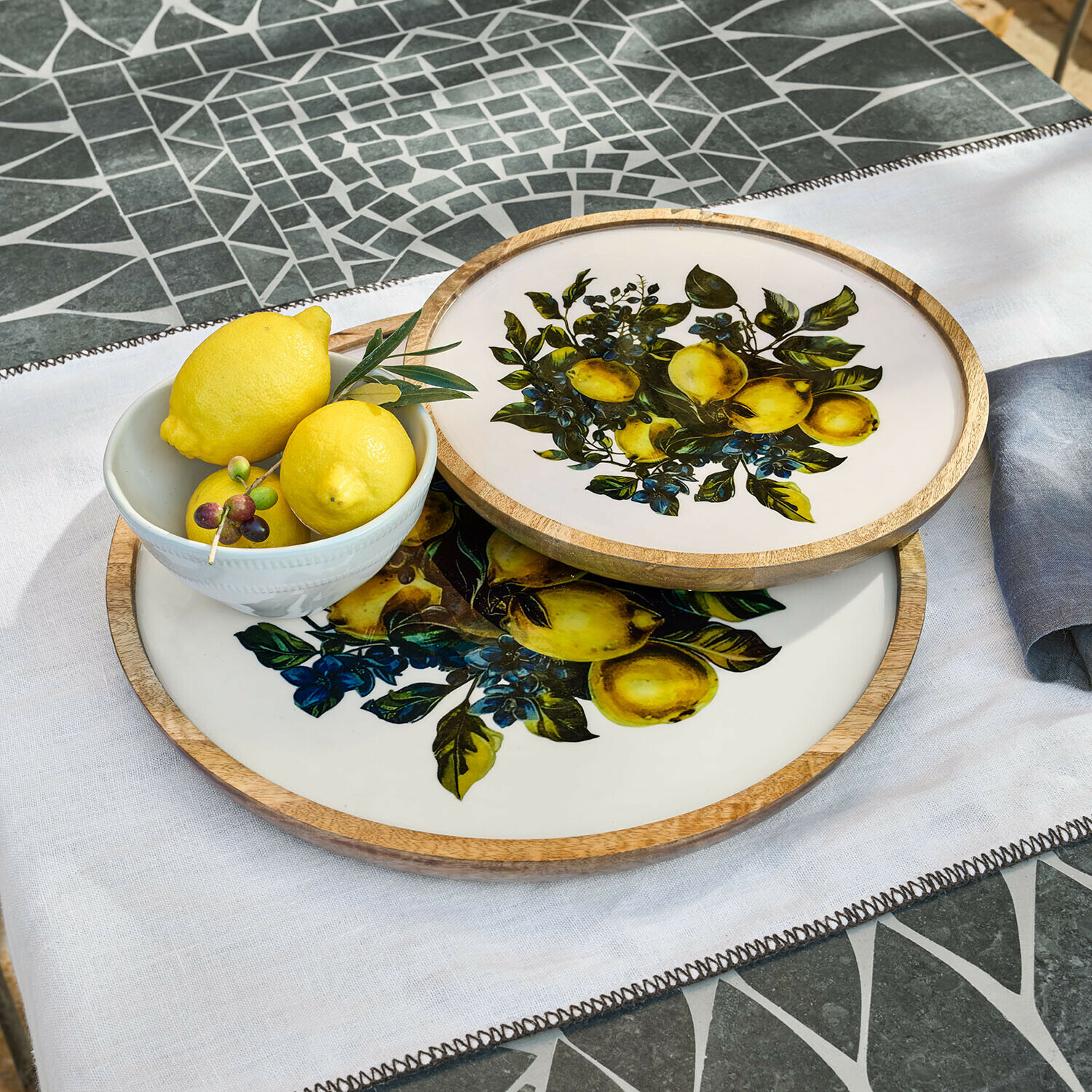 Coppia di vassoi Solianne Due vassoi rotondi in legno e ceramica con motivo limoni su sfondo bianco su tavolo a mosaico